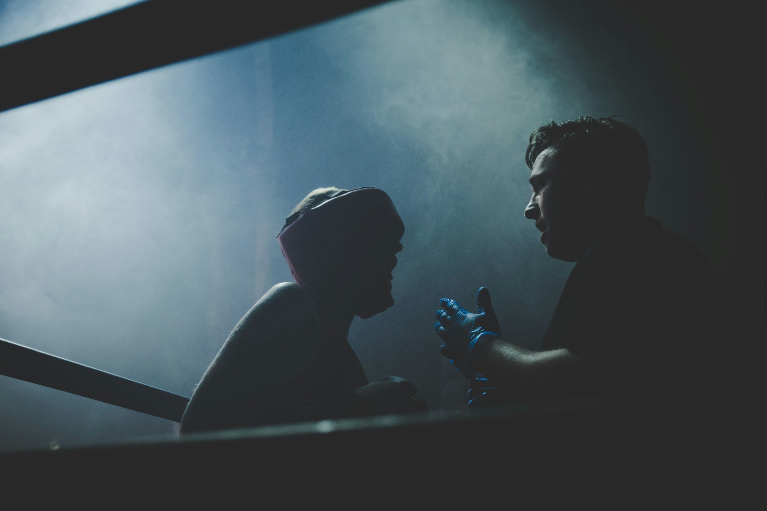 A person wearing gloves and a cap receiving a vaccination shot from another person in a dark, clinical setting with minimal lighting.