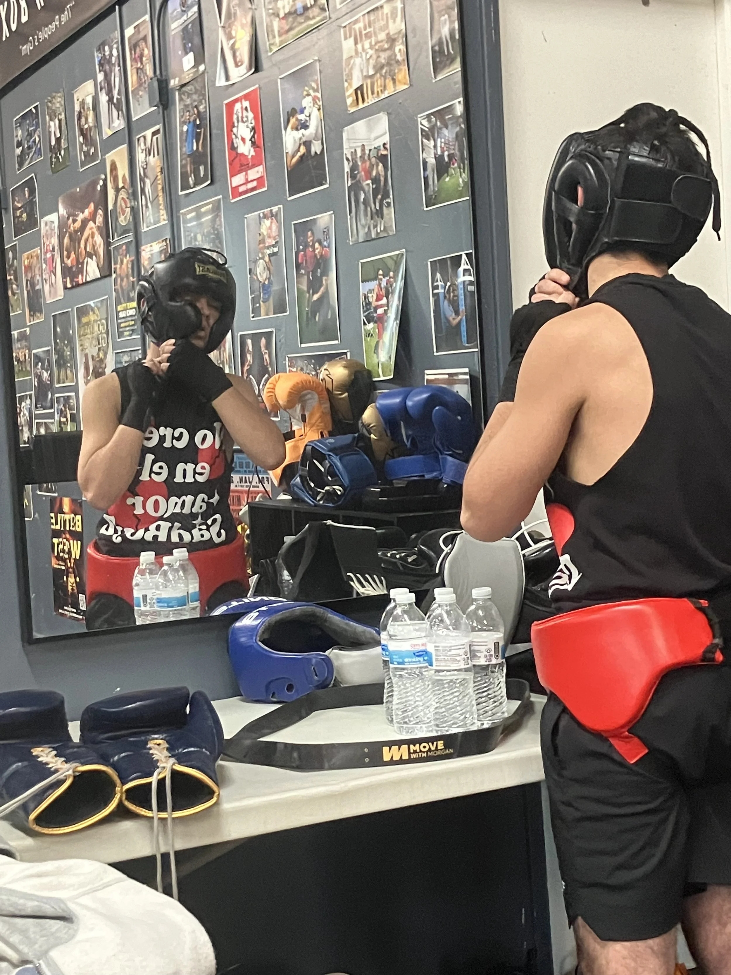 A person preparing for boxing training in a gym, wearing a helmet, gloves, and athletic clothing, with boxing gloves and equipment on the table, and a wall covered with photos of boxing matches and moments.