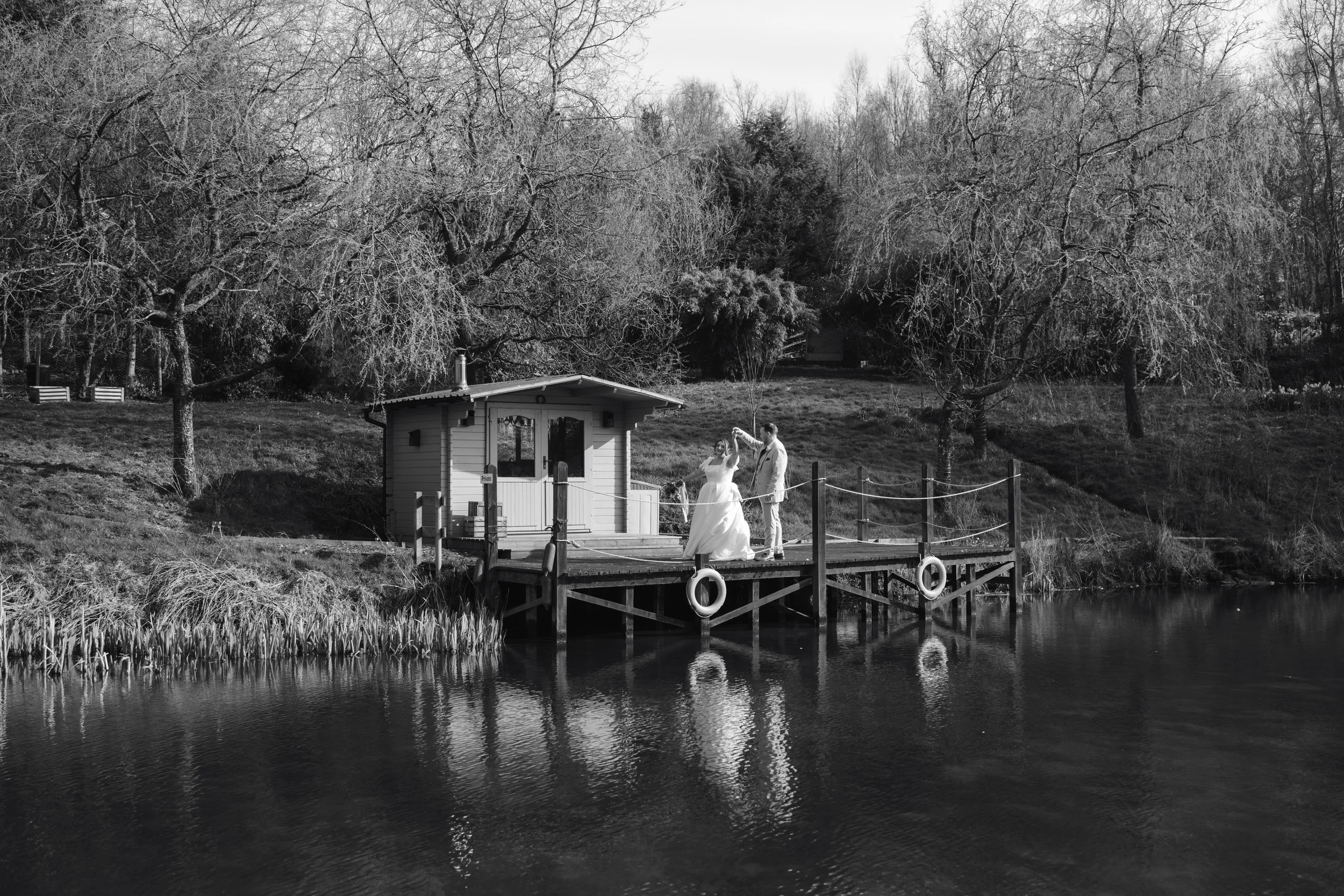 A couple dancing on a small dock near a lake, with a small house behind them and trees with bare branches in the background, in black and white.