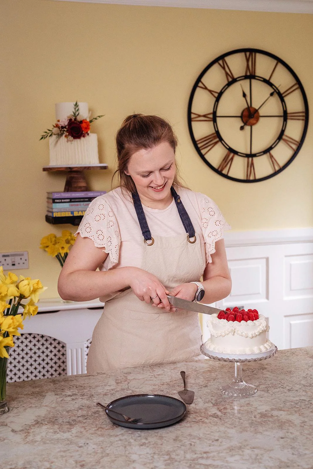 A woman in a beige apron and white shirt is smiling while slicing a wedding cake topped with raspberries in a kitchen.