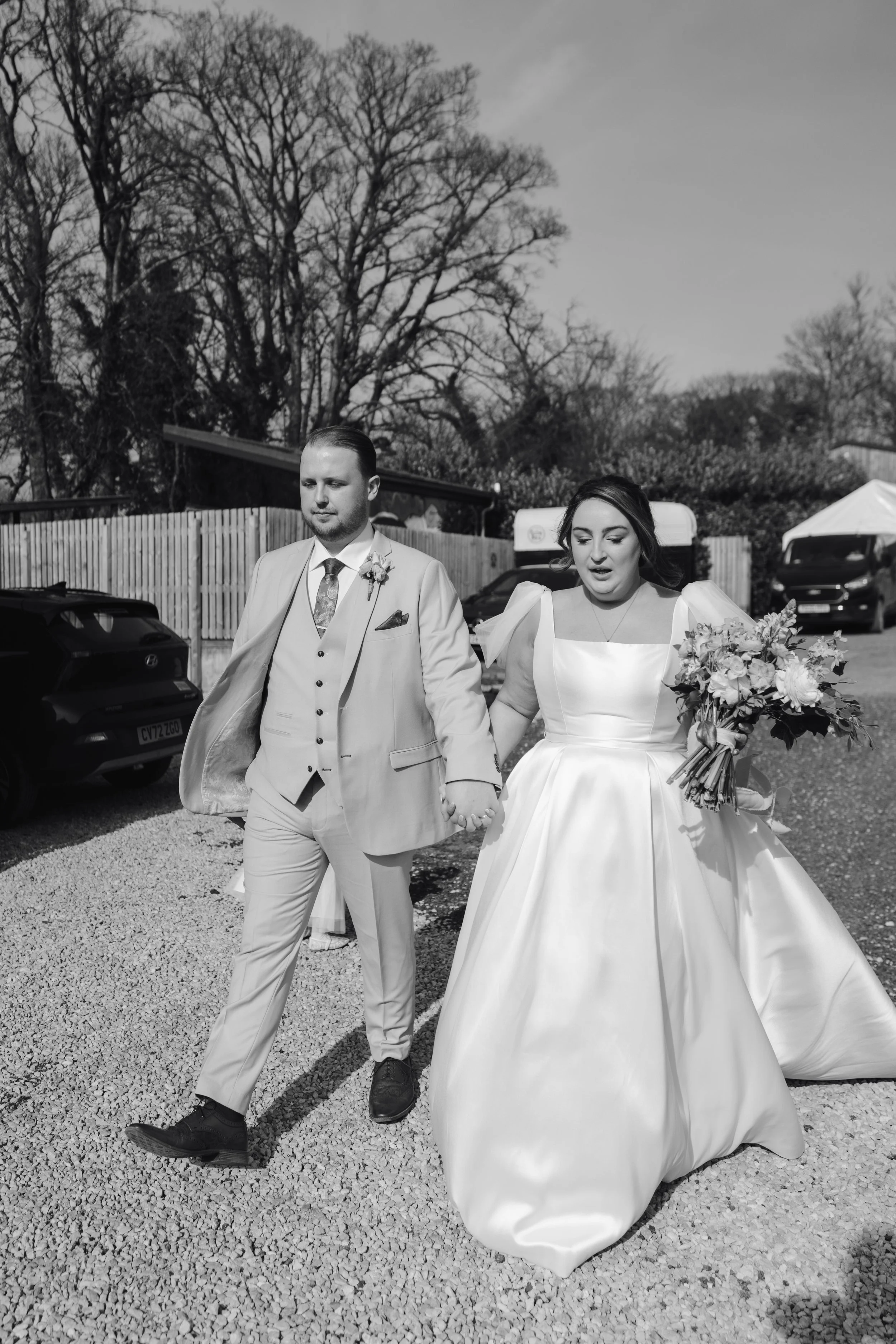 Black and white photo of a bride and groom walking outdoors, holding hands. The bride is wearing a white wedding gown and holding a bouquet, while the groom is dressed in a light-colored suit with a boutonniere. There are cars and a wooden fence in t