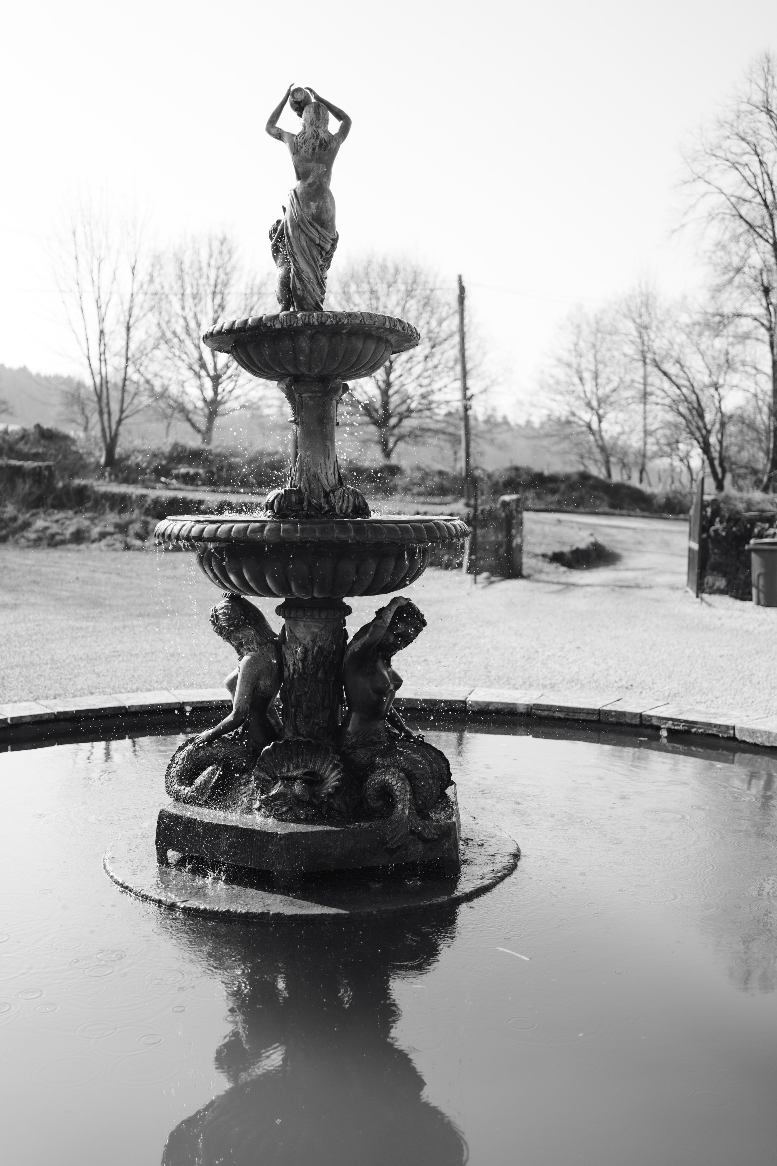 A black and white photograph of an ornate three-tiered fountain with cherub sculptures, water flowing from the top, and a reflection in the water basin below. Trees and a clear sky are visible in the background.