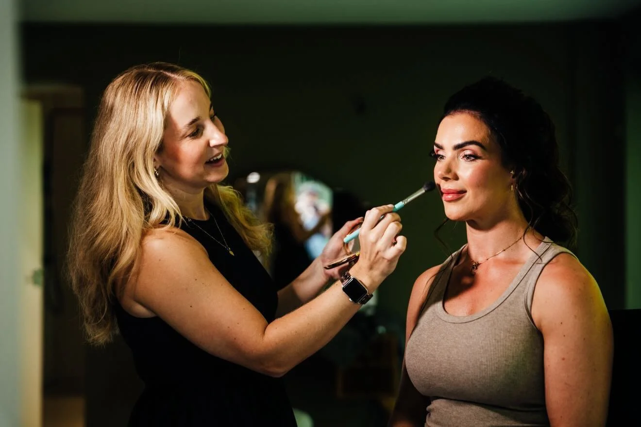 A makeup artist applying makeup to a woman in a sleeveless top in a salon.