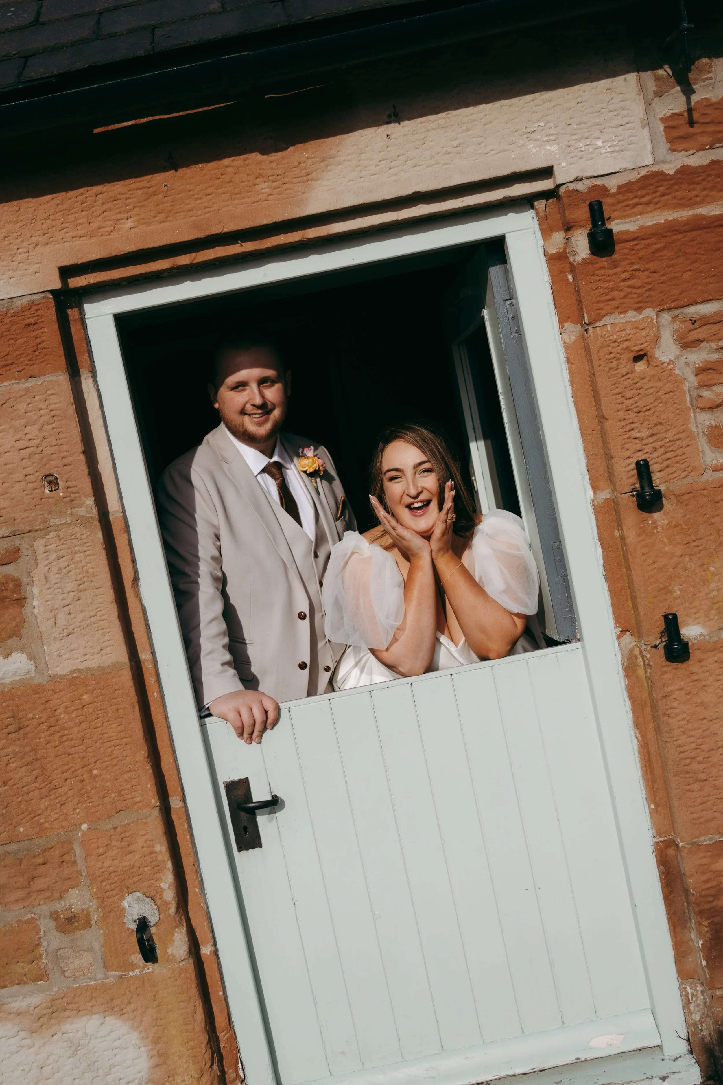 A man in a beige suit and a woman in a white dress looking out a small window in a brick building, smiling and excited.