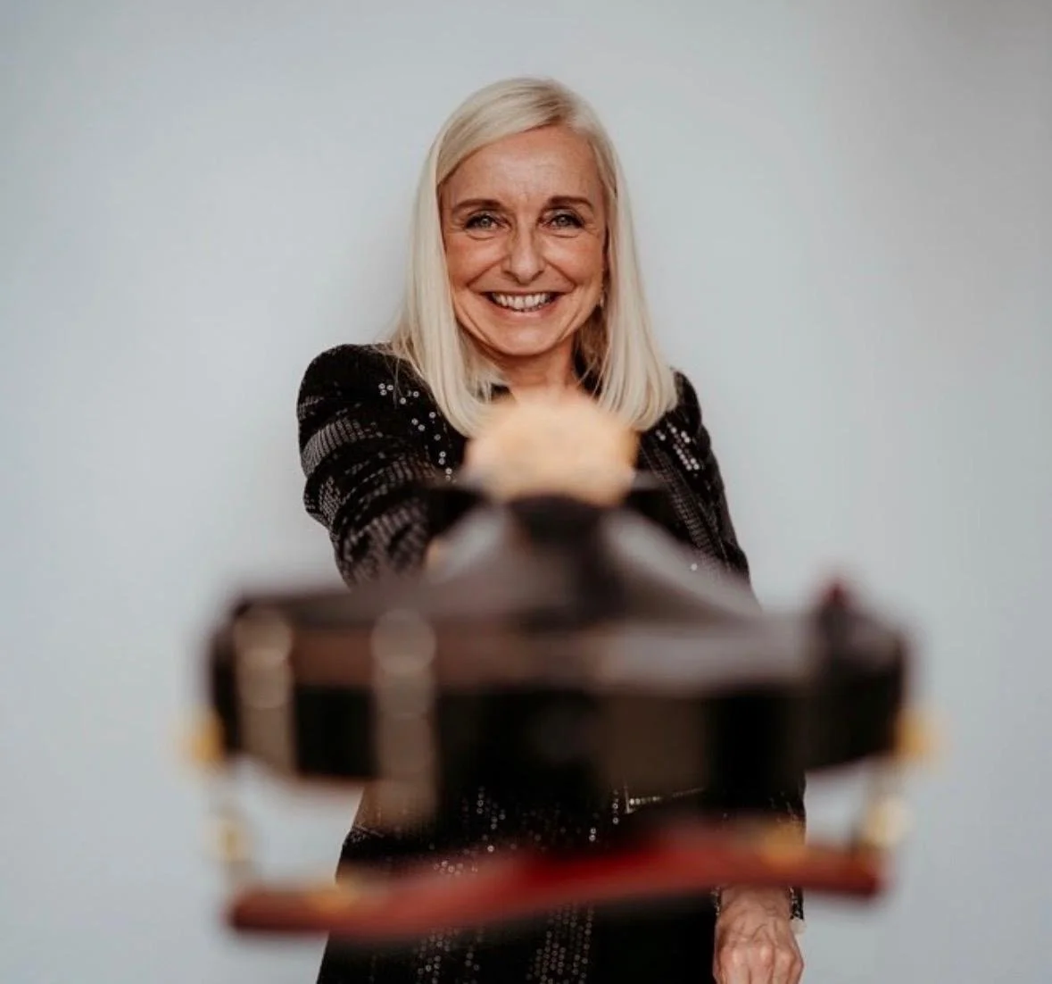 A woman with blonde hair smiling at the camera, holding a violin in front of her against a plain white background.