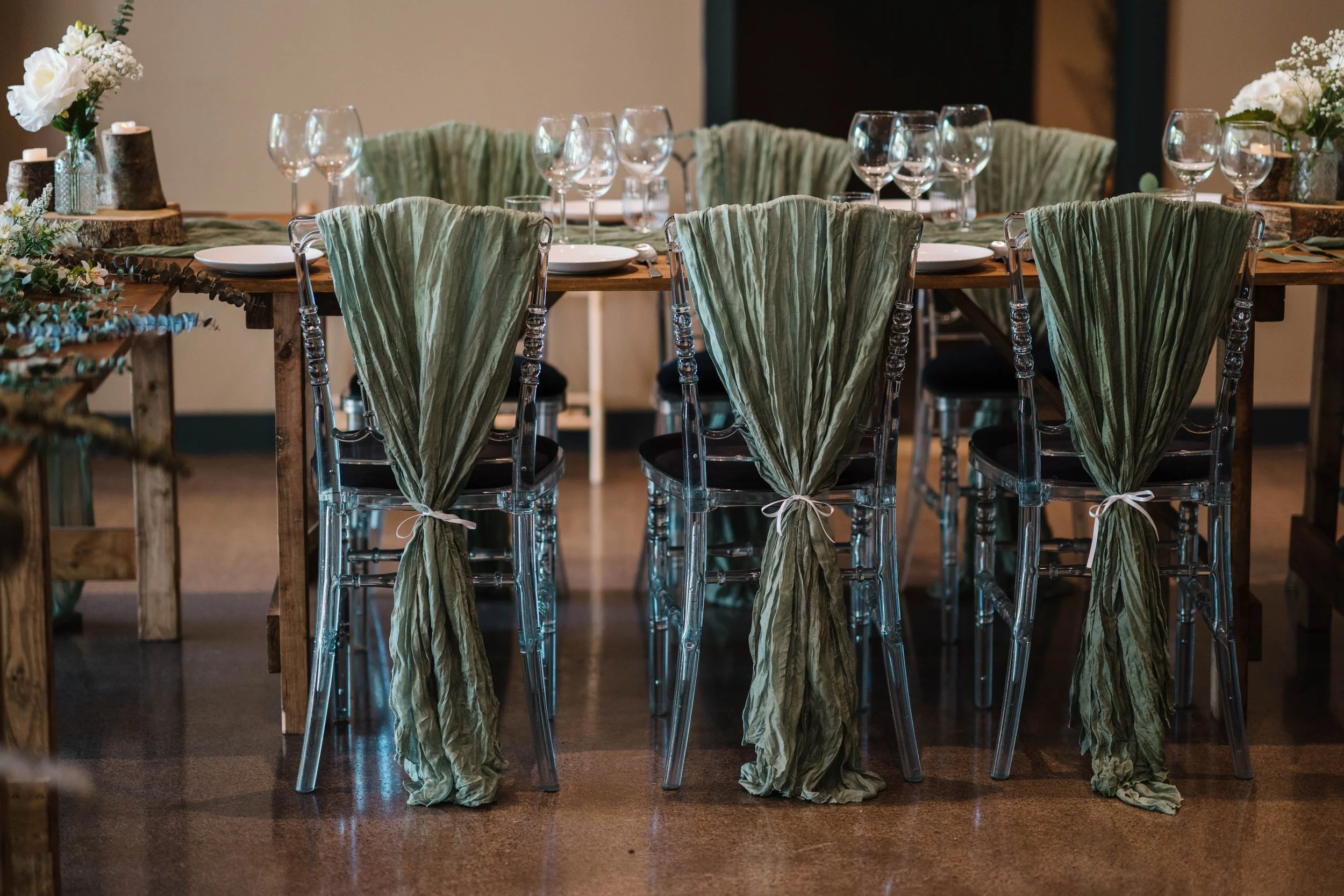 A rustic dining table set with white plates, wine glasses, and green tied chair covers, decorated with floral arrangements in vases and wood slices.