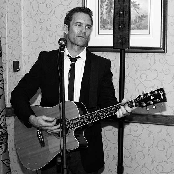 A man in a suit playing an acoustic guitar and singing into a microphone in a decorated room.