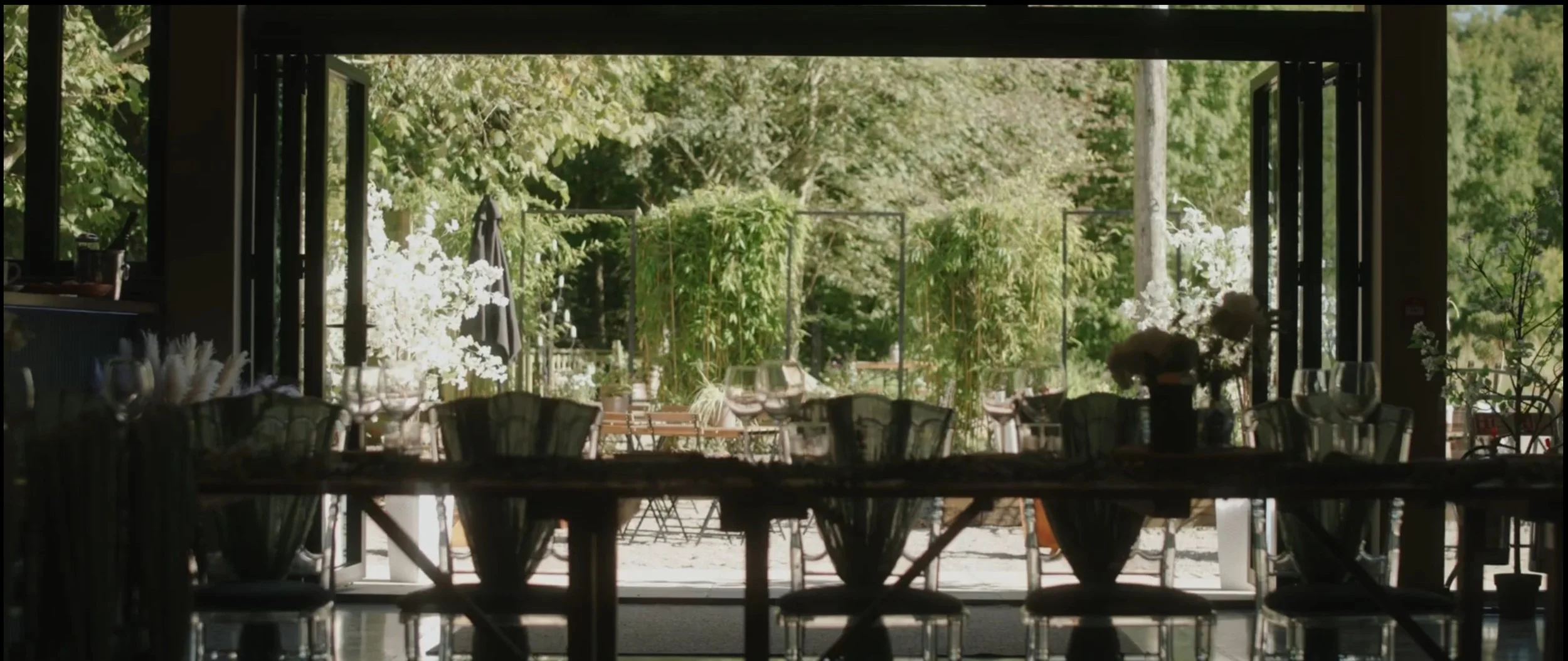 View of a restaurant dining area through large open windows, decorated with flowers and greenery outside.