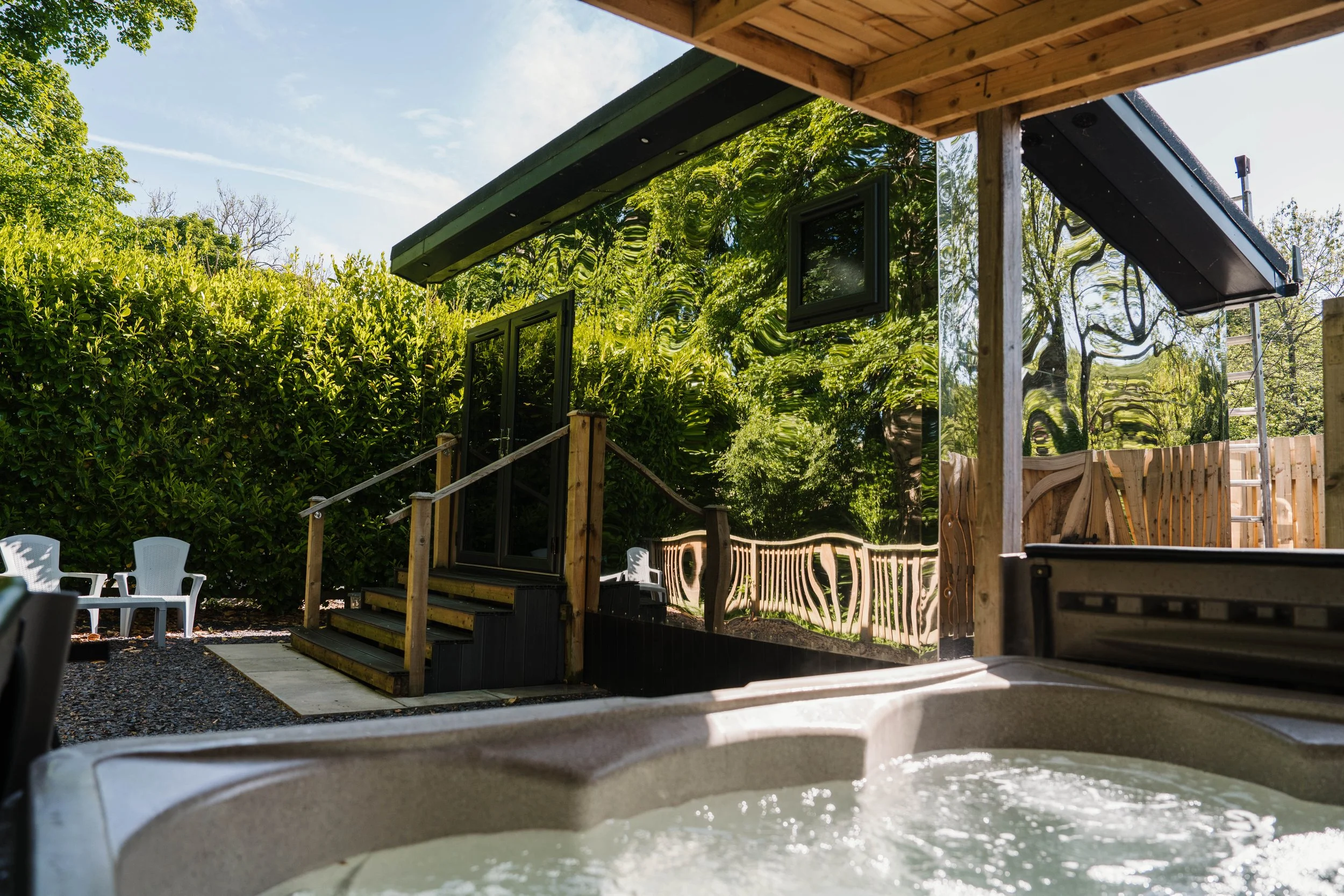 A hot tub with water, viewed from inside, overlooking a backyard with a wooden deck, chairs, lush green trees, and a house with a reflective black exterior wall.