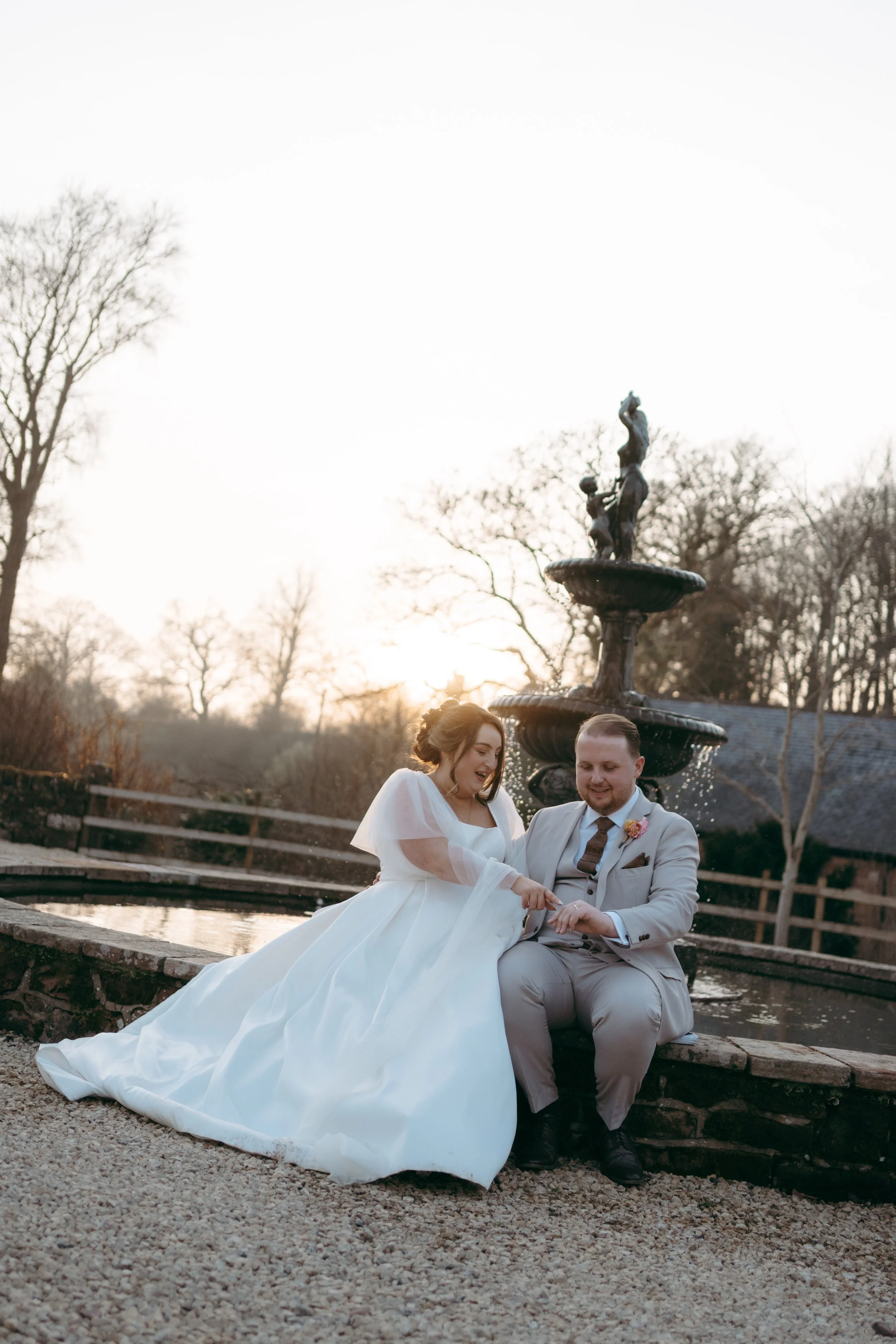 A newlywed couple in wedding attire sitting by a fountain in an outdoor garden, holding hands and smiling at each other during sunset.