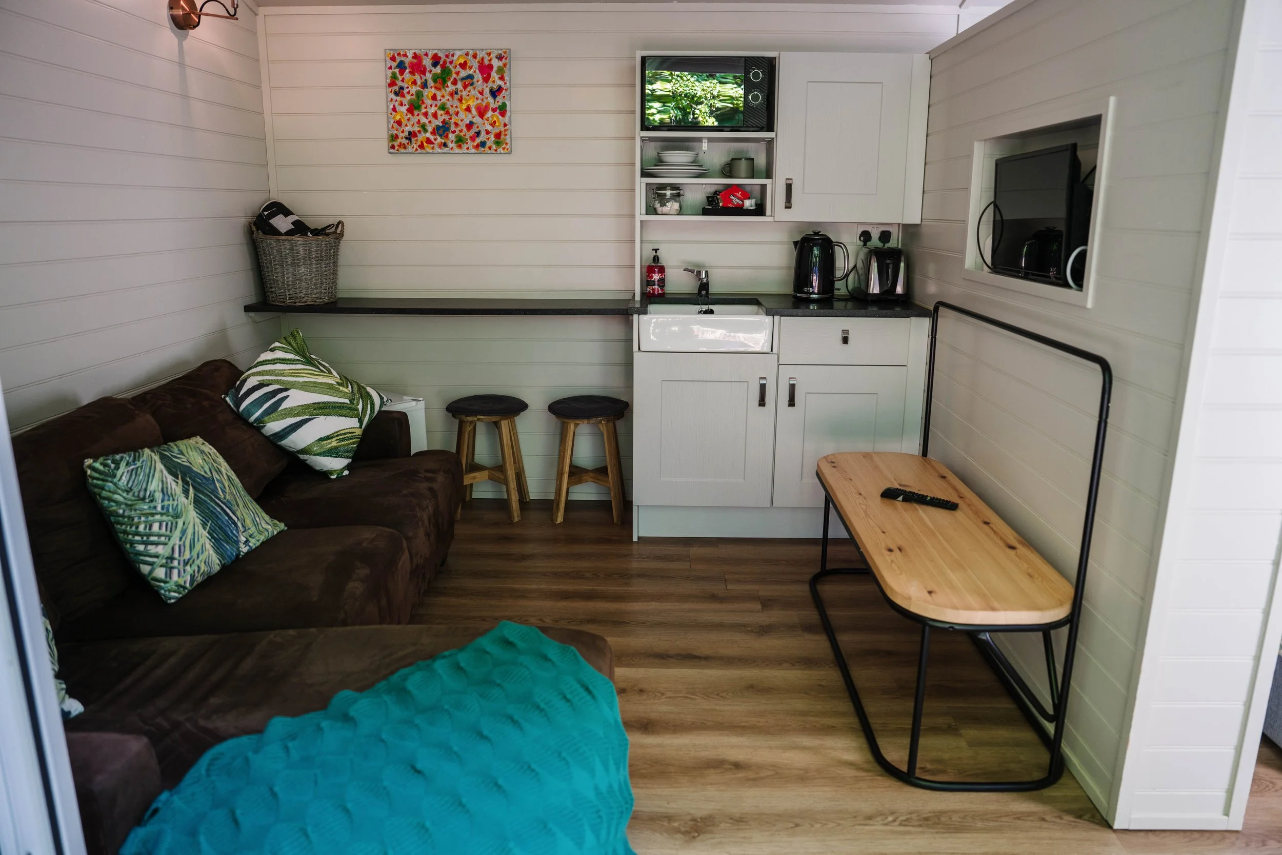 Small living space with a brown sofa decorated with tropical leaf patterned pillows, a wall-mounted artwork, a kitchenette with a sink, kettle, and toasters, two wooden bar stools, and a wooden table with a TV remote on it.