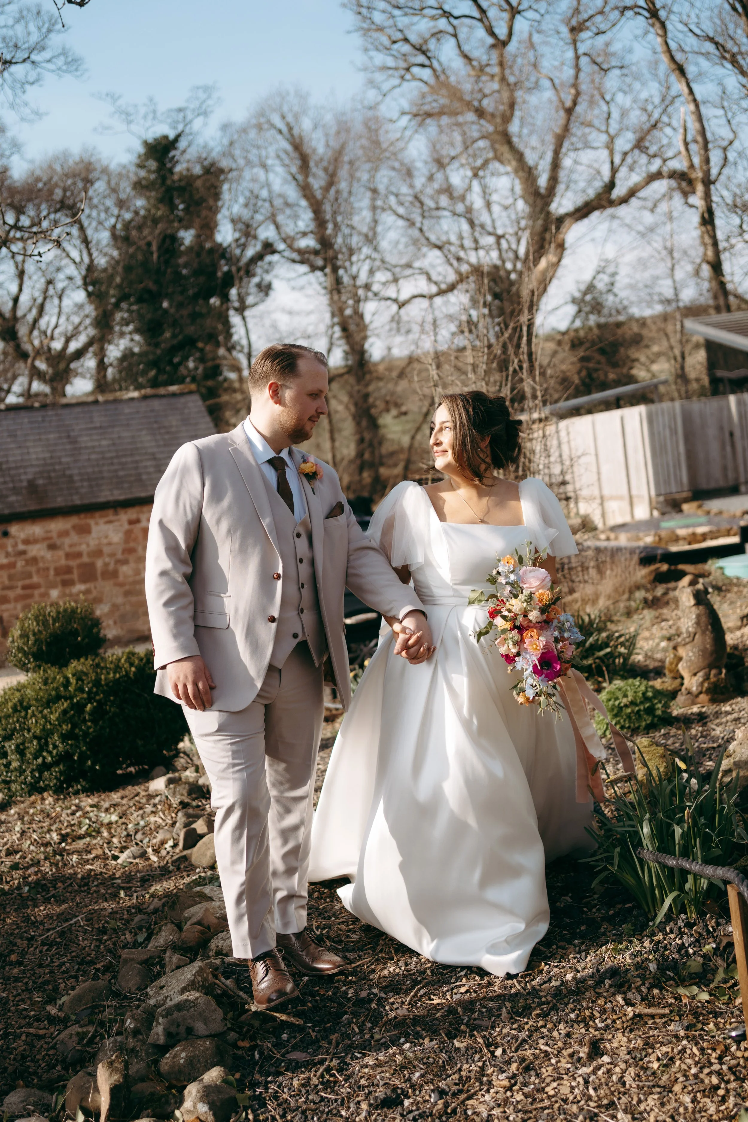 A newlywed couple walking outdoors in a garden, holding hands, with bare trees and a wooden fence in the background.