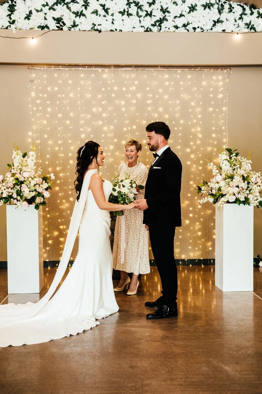 A wedding ceremony with a bride and groom holding hands, smiling at each other, in front of an elegant backdrop of string lights and floral arrangements, with an officiant standing behind them.