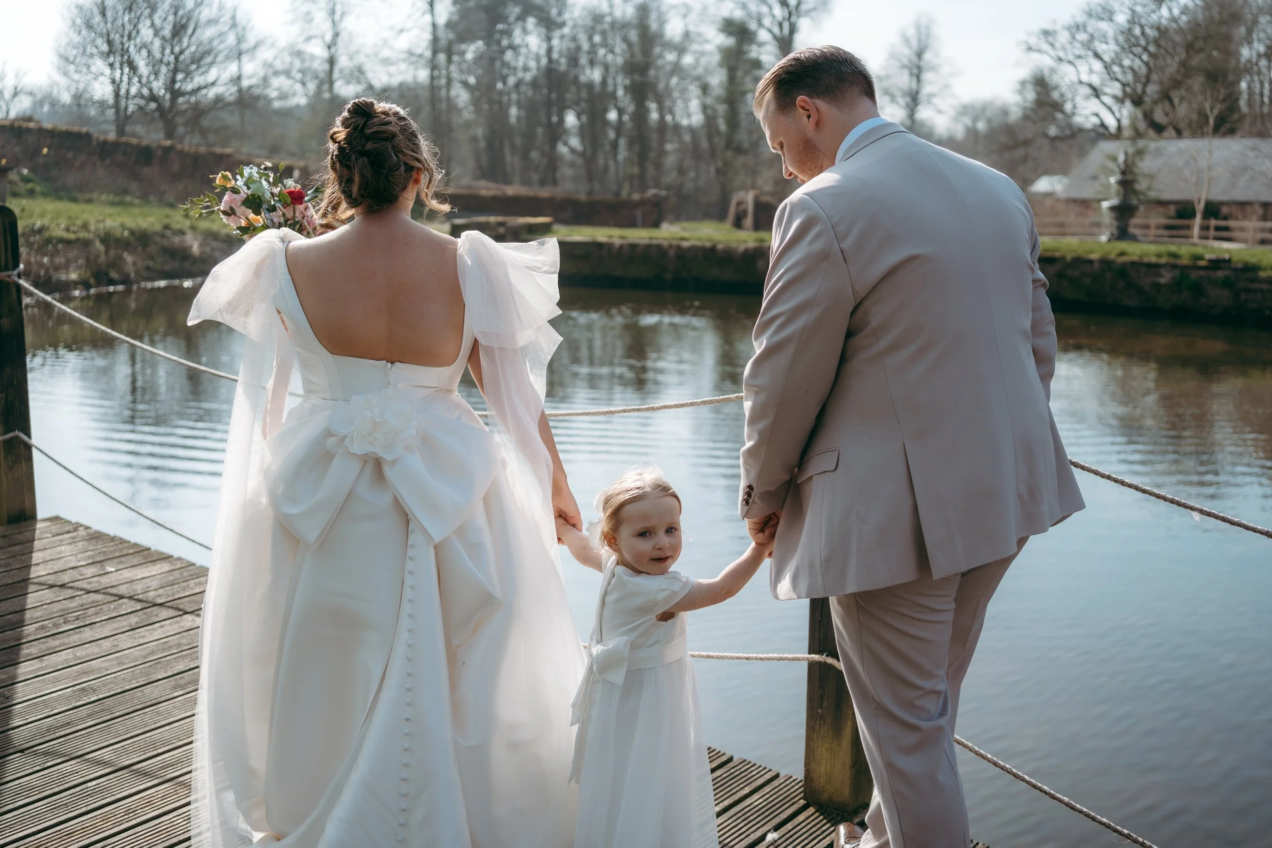 A couple, a woman in a white wedding dress holding a bouquet and a man in a light gray suit, holding hands with two young girls in white dresses, standing on a wooden dock next to a body of water, with trees and buildings in the background on a clear