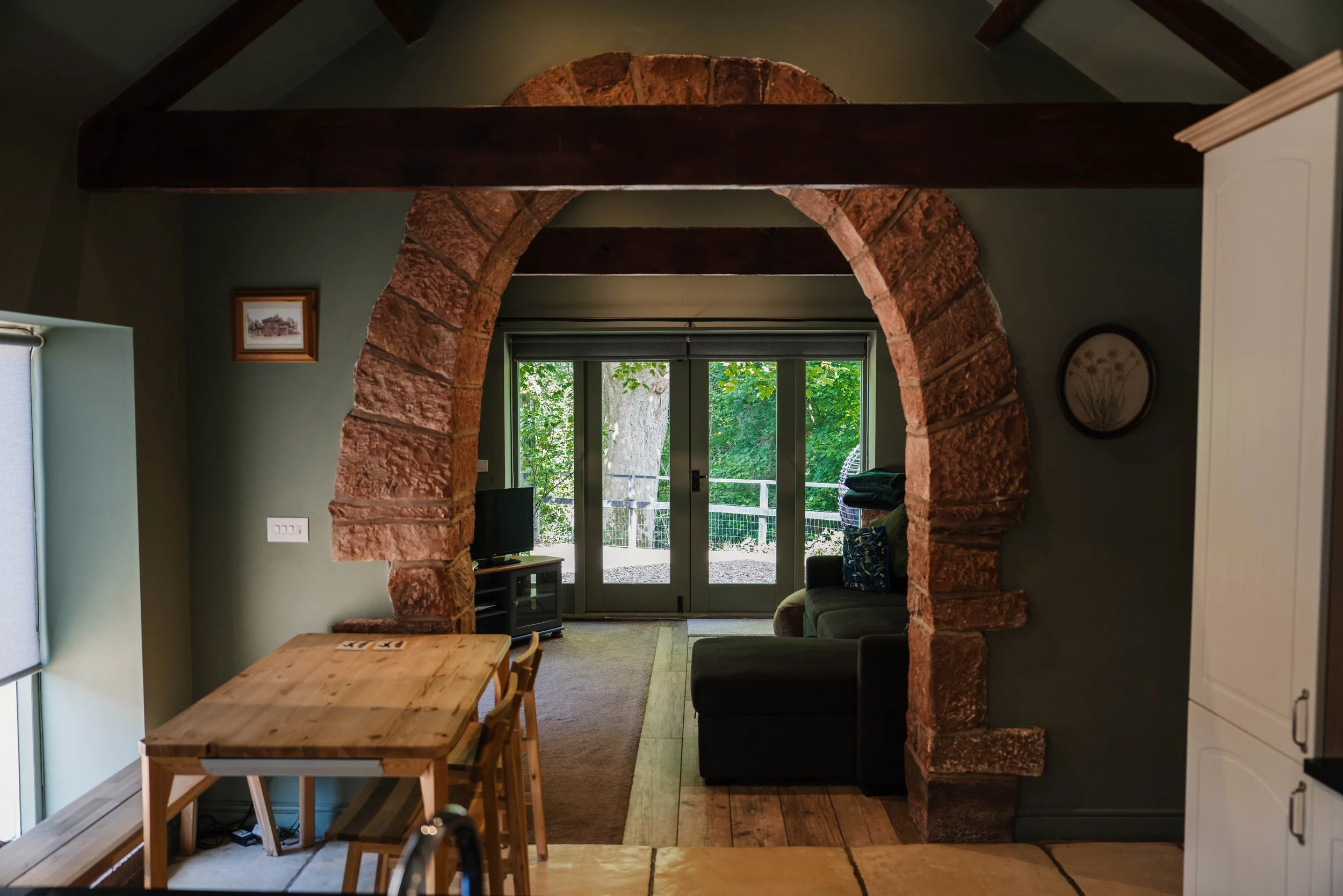 Living room featuring a brick archway, large windows showing greenery outside, a small wooden table with chairs, a gray armchair, a small TV, cabinet, and decorative wall art.
