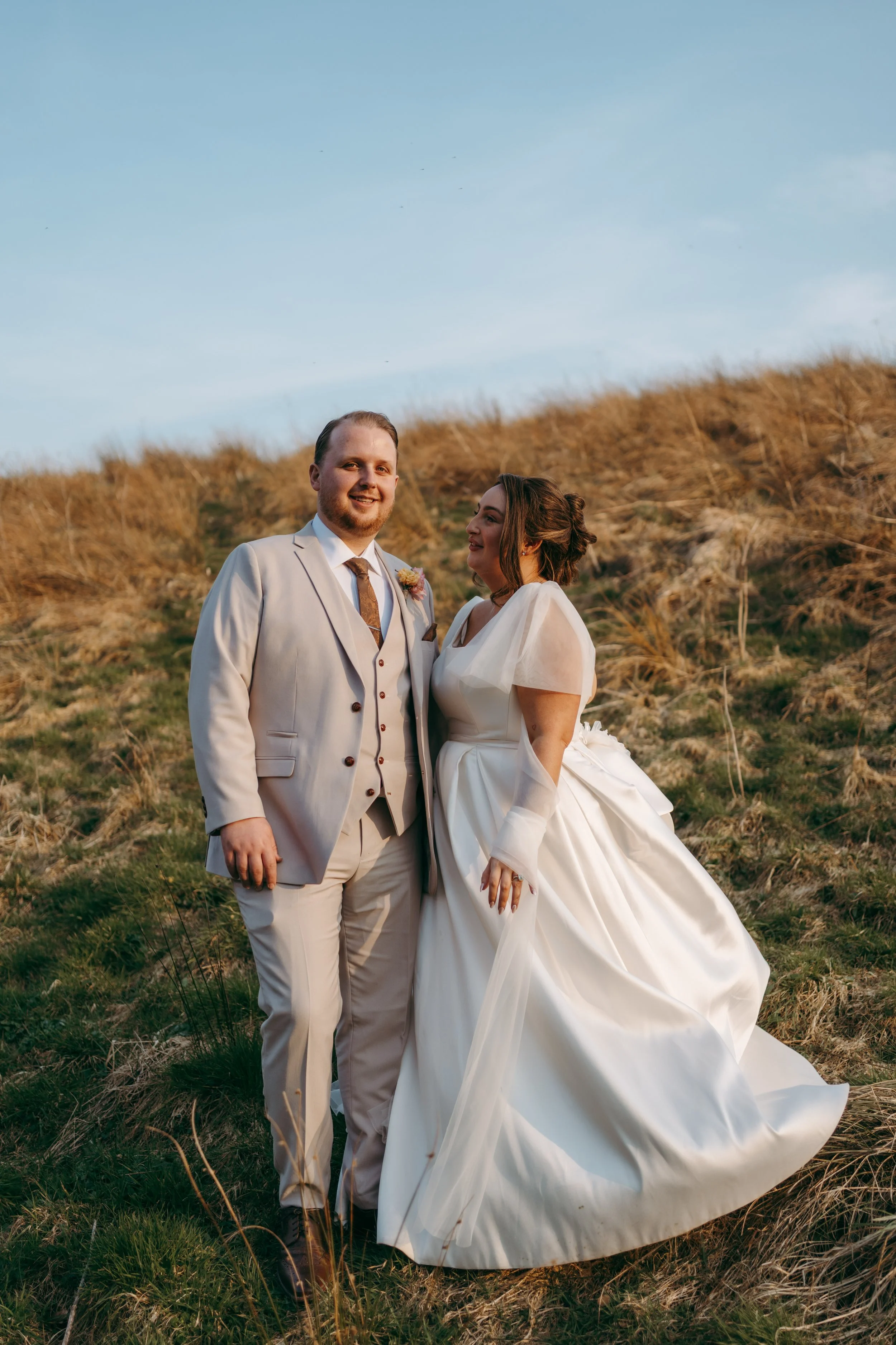 A bride and groom standing outdoors with hills in the background, dressed in wedding attire, smiling and looking at each other during sunset.