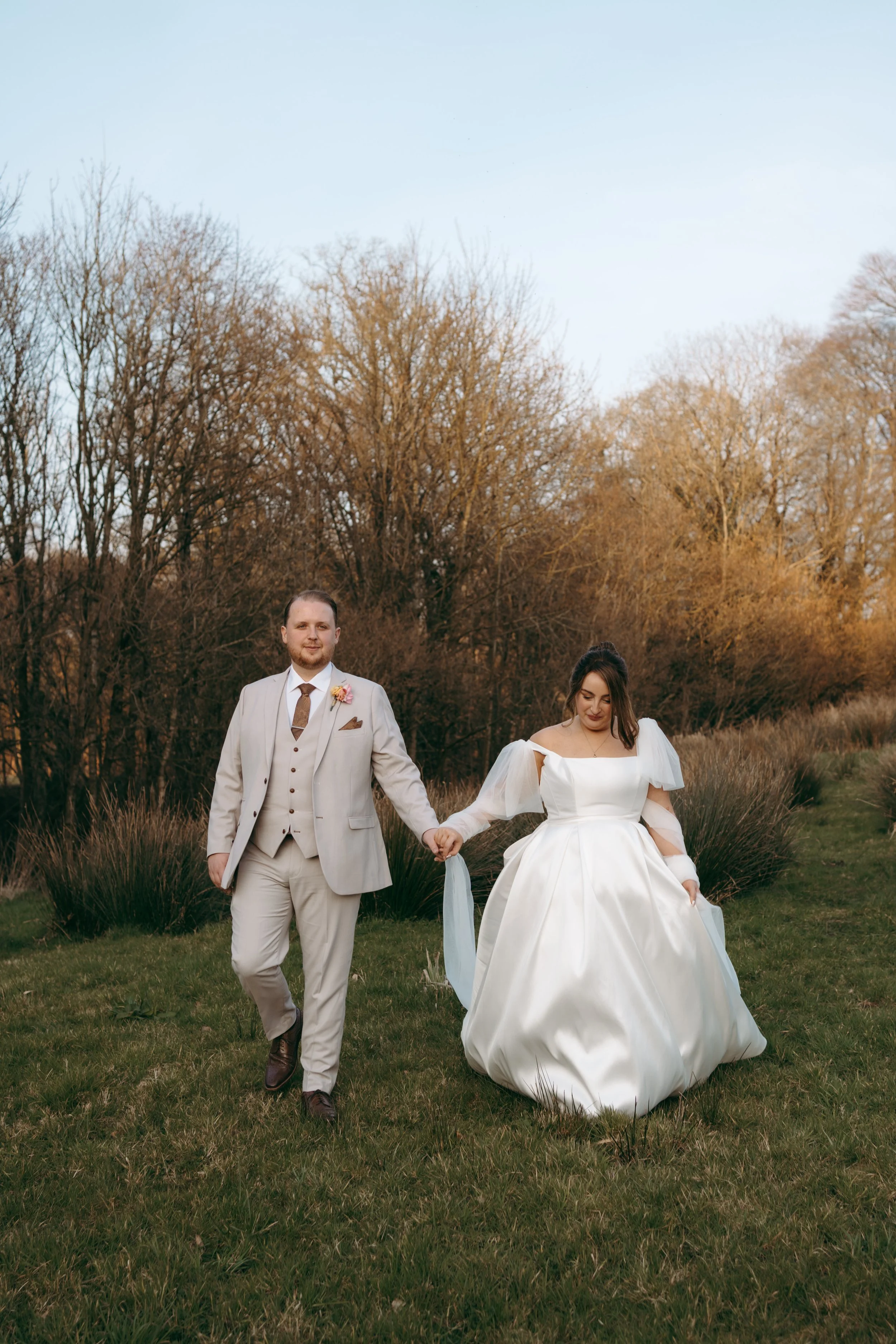 A couple in wedding attire walking hand in hand in a grassy outdoor area with trees and a clear blue sky in the background.
