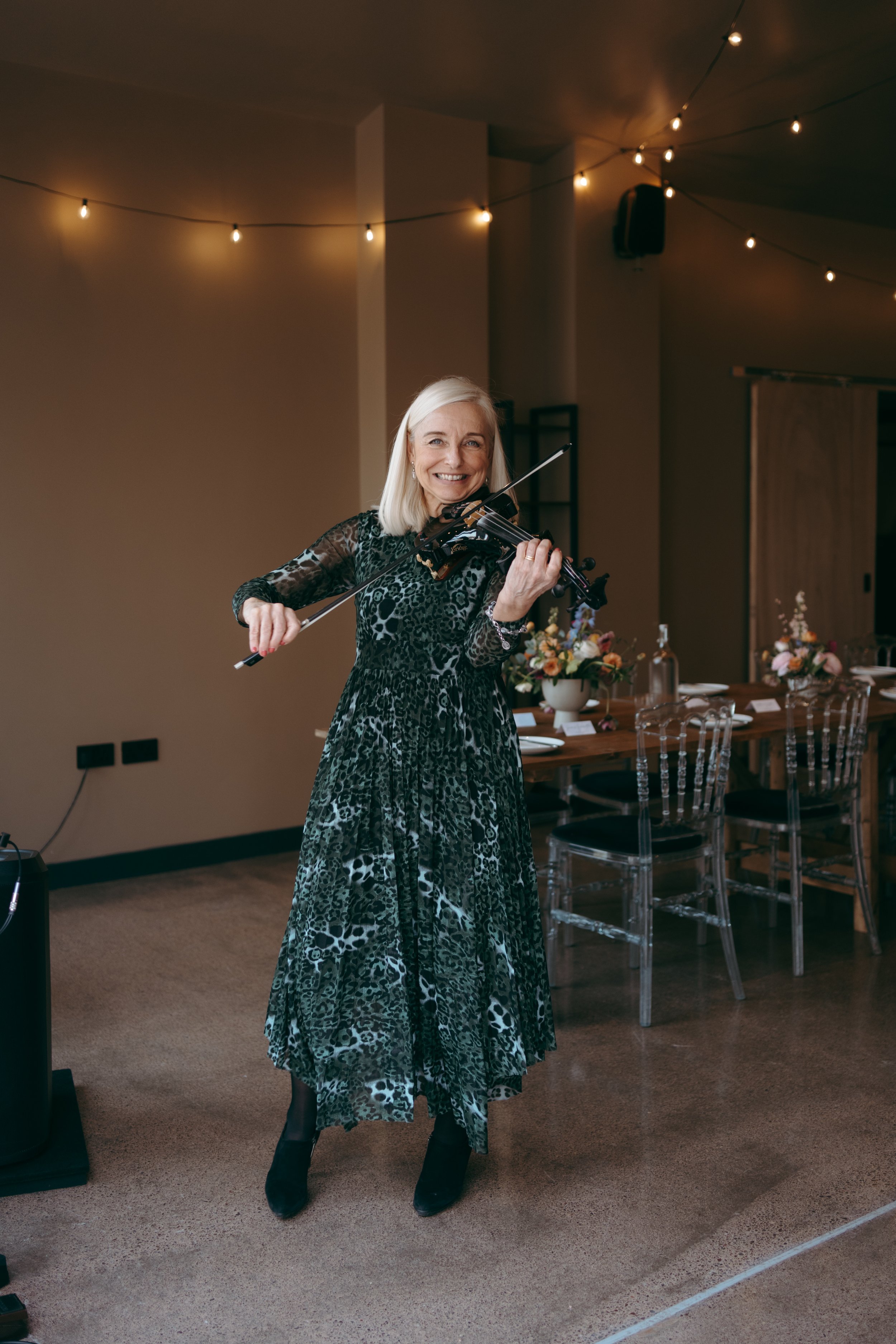 A woman with blonde hair is playing a violin and smiling at an indoor event with string lights and a decorated table with flowers.