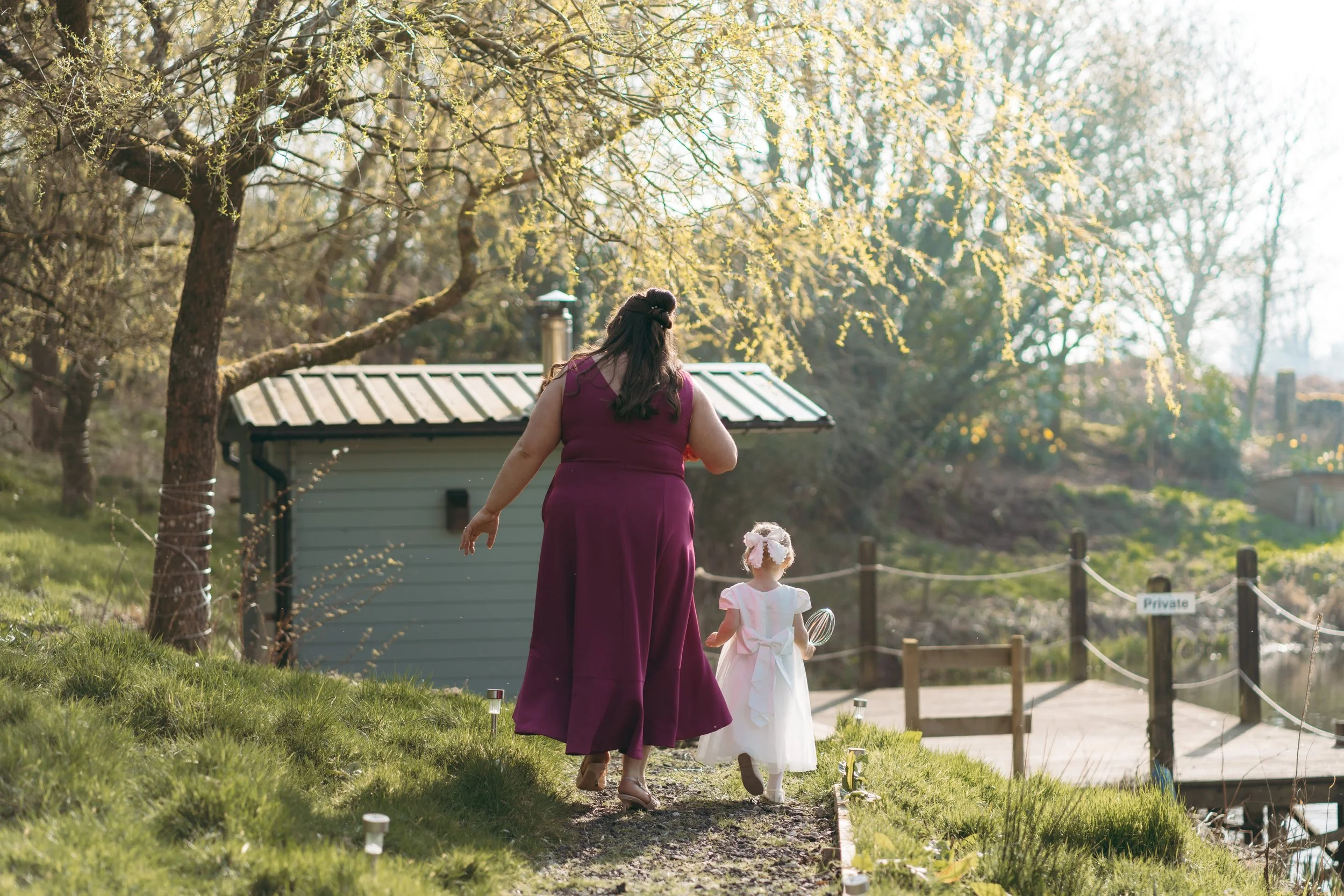 A woman in a purple dress and a young girl in a light pink dress walking on a grassy area near a small gray building and trees.
