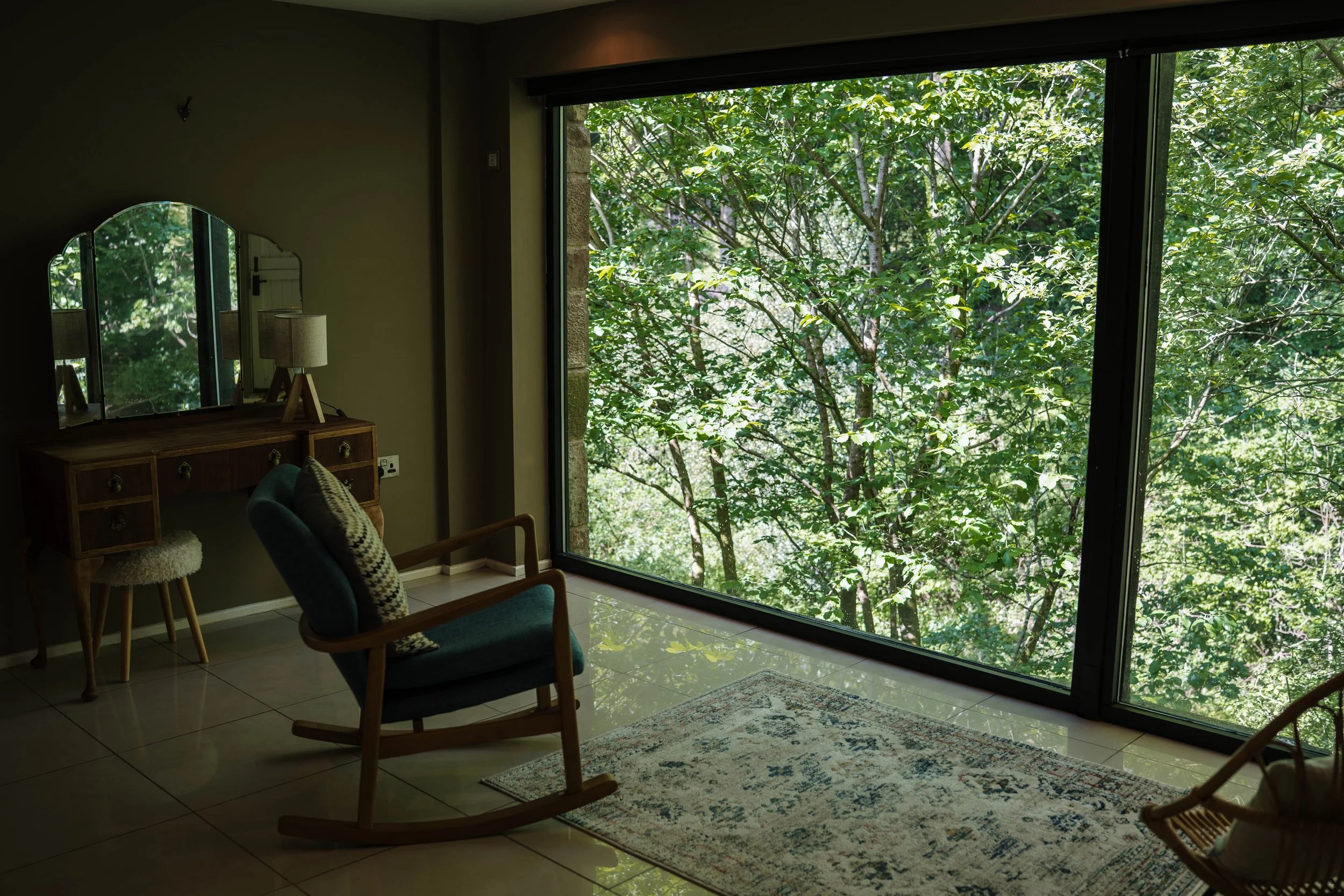 Interior of a room with a large window showing a dense green forest outside, a vintage wooden dresser with a three-part mirror, a table lamp, a patterned chair, and a colorful rug on a tiled floor.