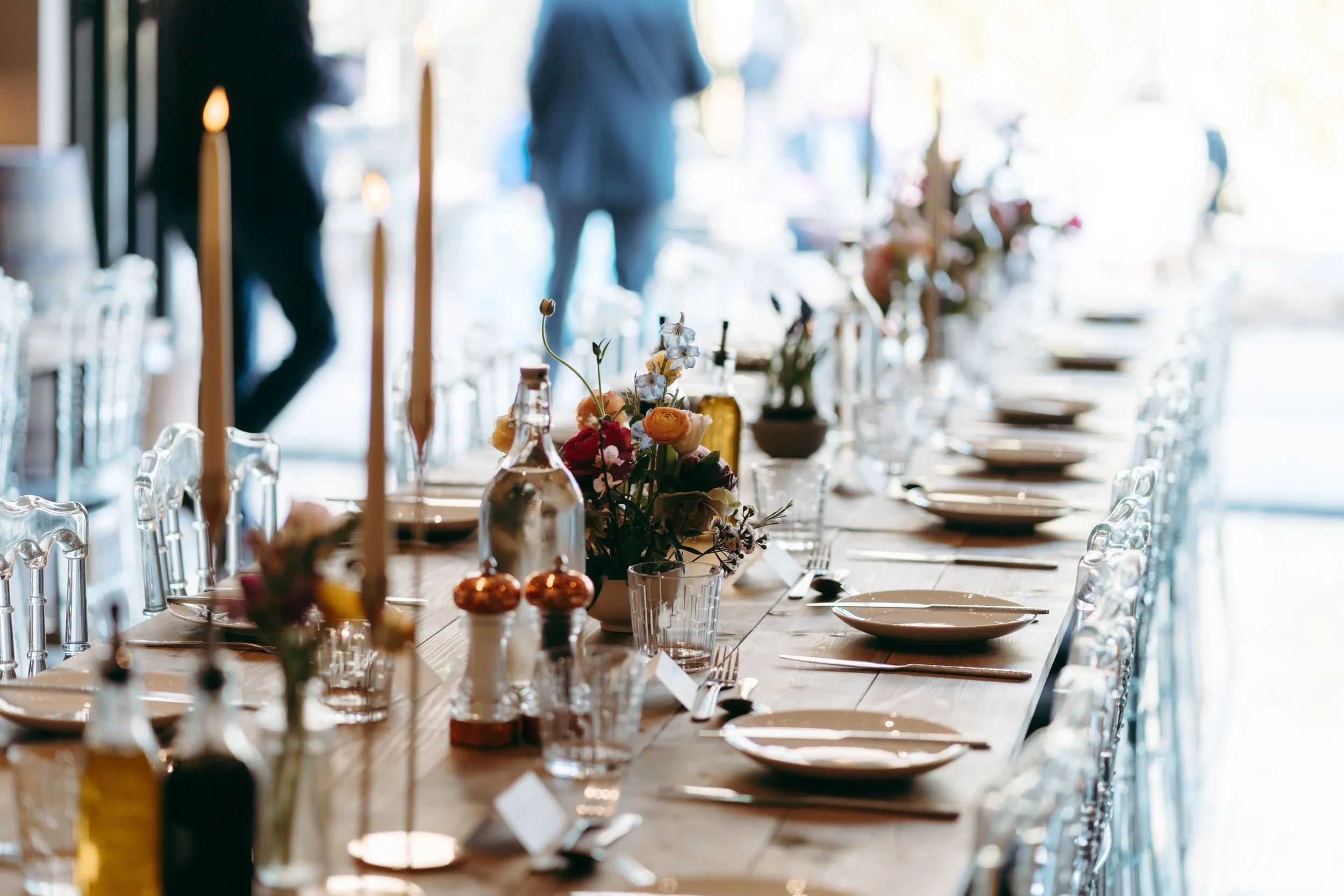 A long dining table set for a gathering or event with plates, glasses, cutlery, and floral centerpieces, in a well-lit venue with people standing in the background.