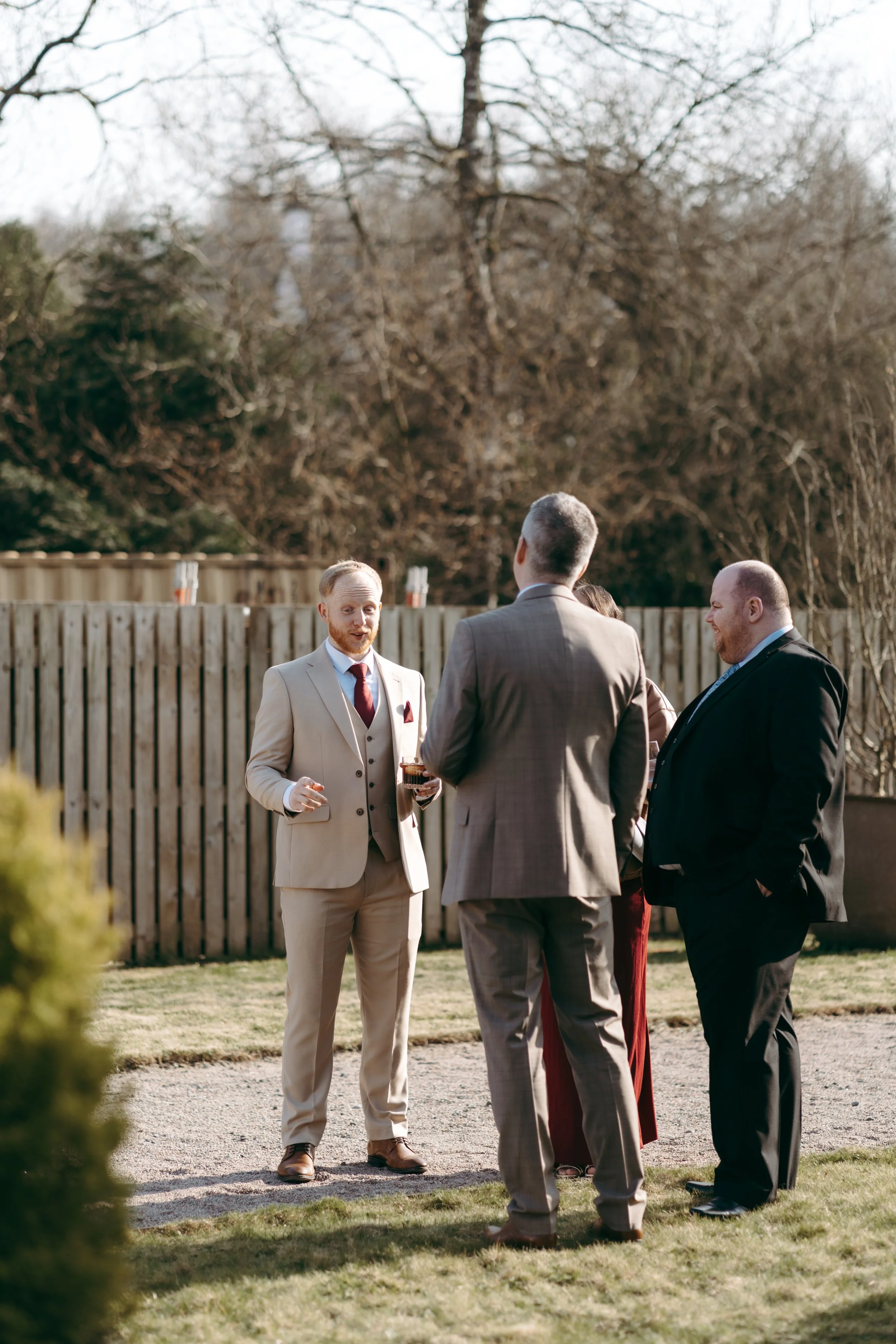 Group of four men in suits talking outdoors in a backyard with a wooden fence and bare trees, sunny day.