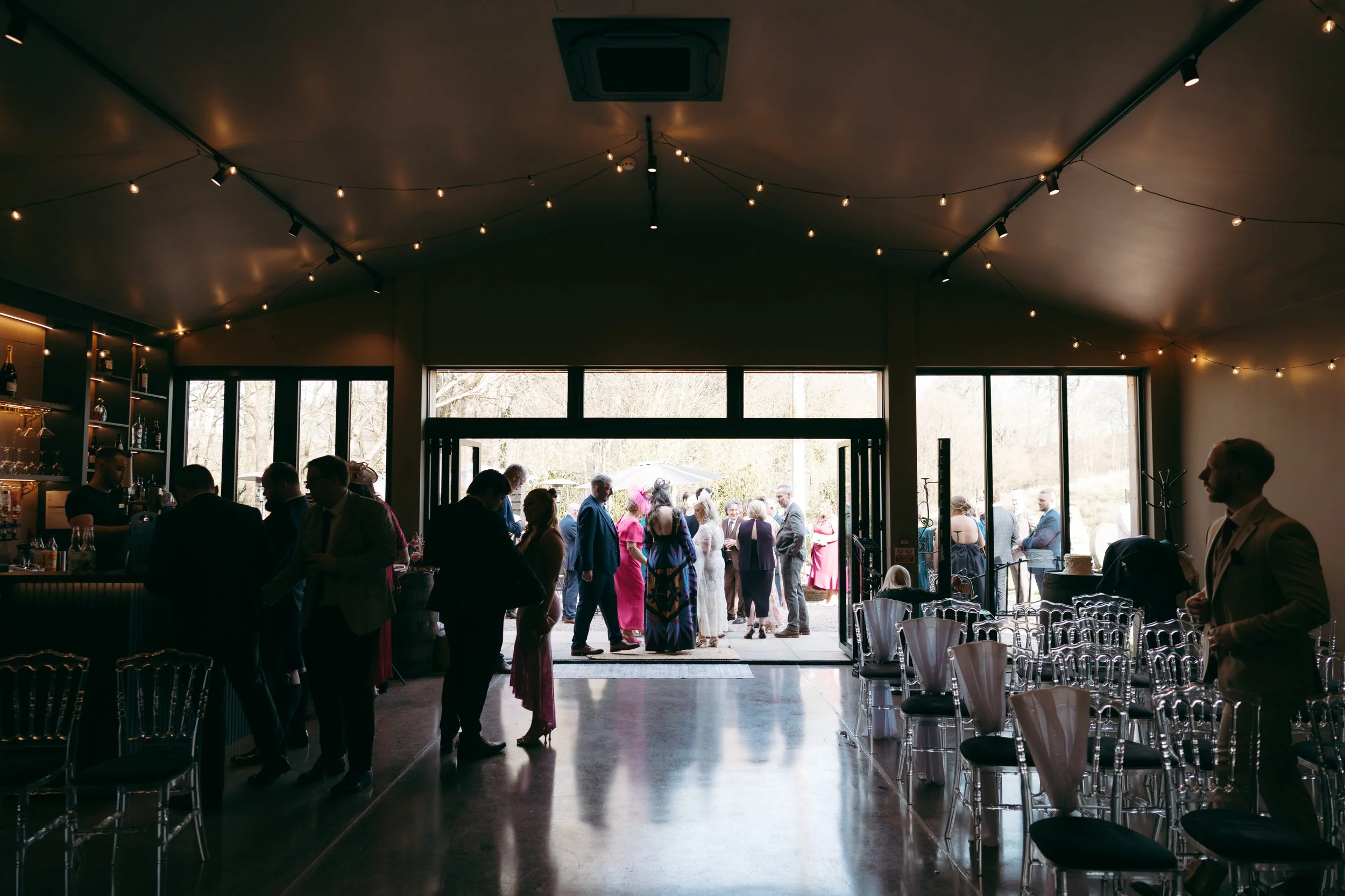 People gathered at the entrance of a venue with large windows and glass doors, outside seen through the doors are trees and a cloudy sky. Inside is decorated with string lights hanging from the ceiling, and some people are sitting while others are st