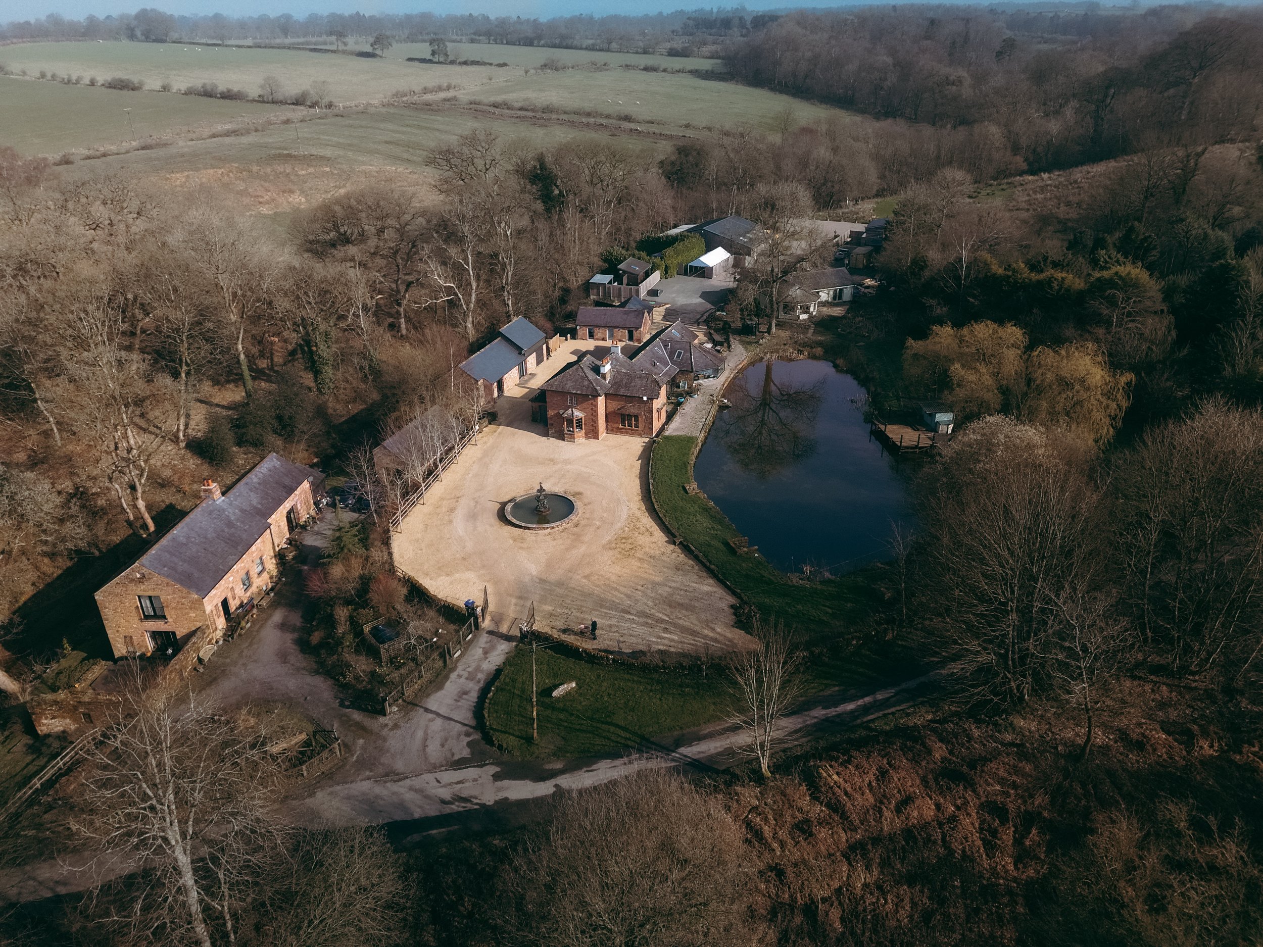 Aerial view of a residential property surrounded by trees and open land, featuring a pond, farm buildings, and a large house with a circular driveway and a fountain.