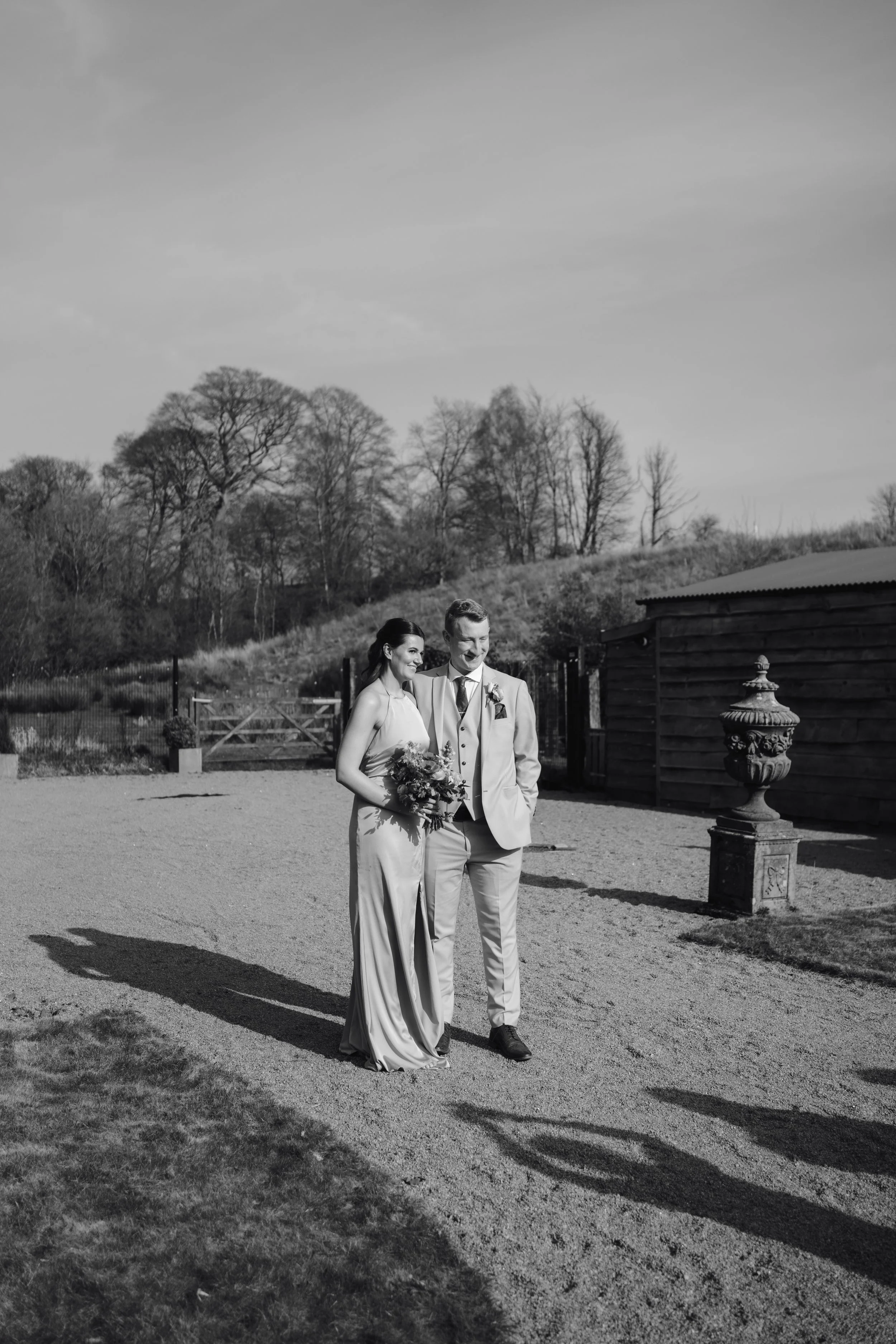 A black and white photograph of a bride and groom standing outdoors on a sunny day, holding hands and smiling. The bride is wearing a long gown and holding a bouquet, while the groom is in a light-colored suit with a boutonniere. They are surrounded 