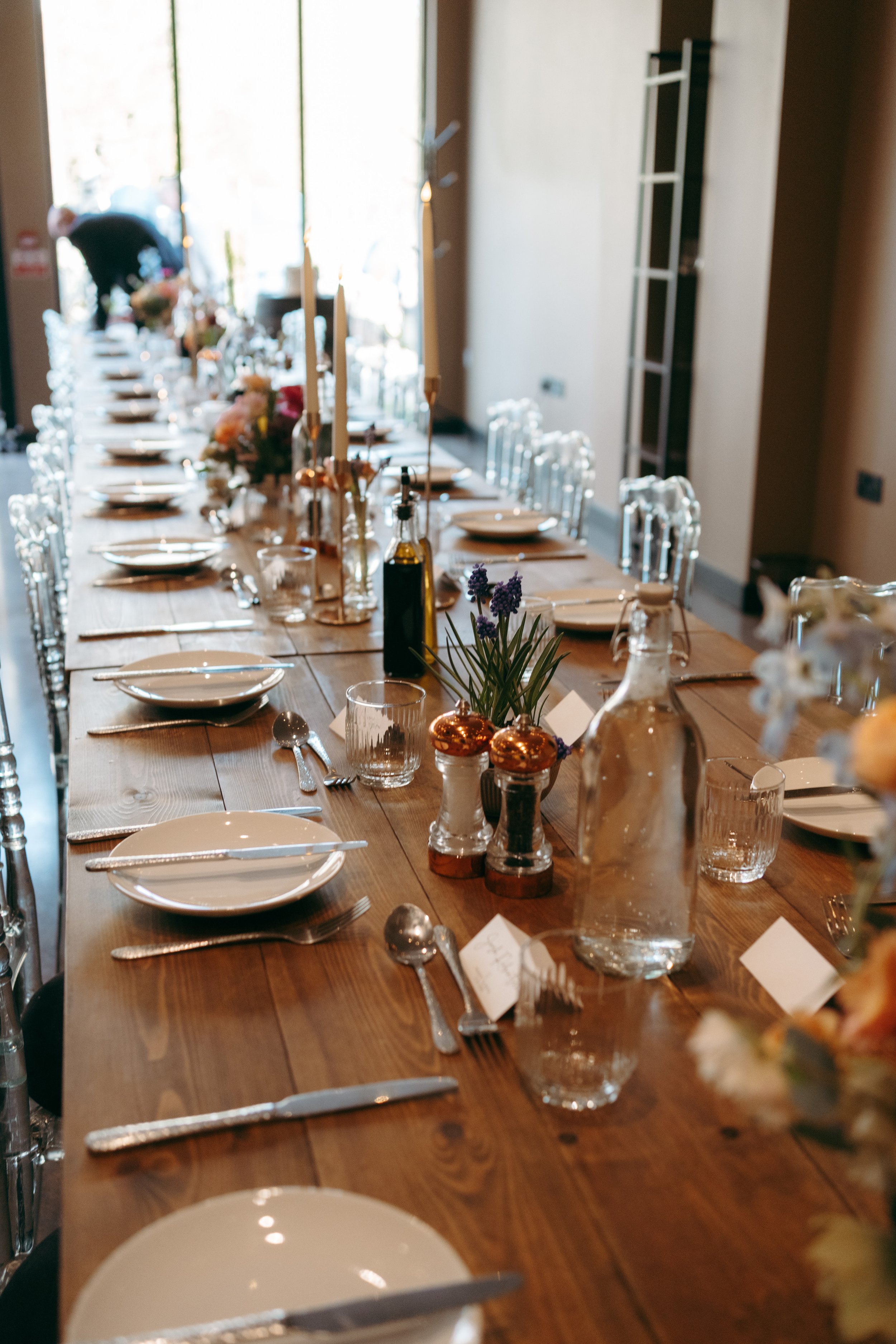 A long wooden dining table set for a meal with white plates, silverware, glasses, and decorated with flowers, candles, and vases, inside a room with a large window in the background.