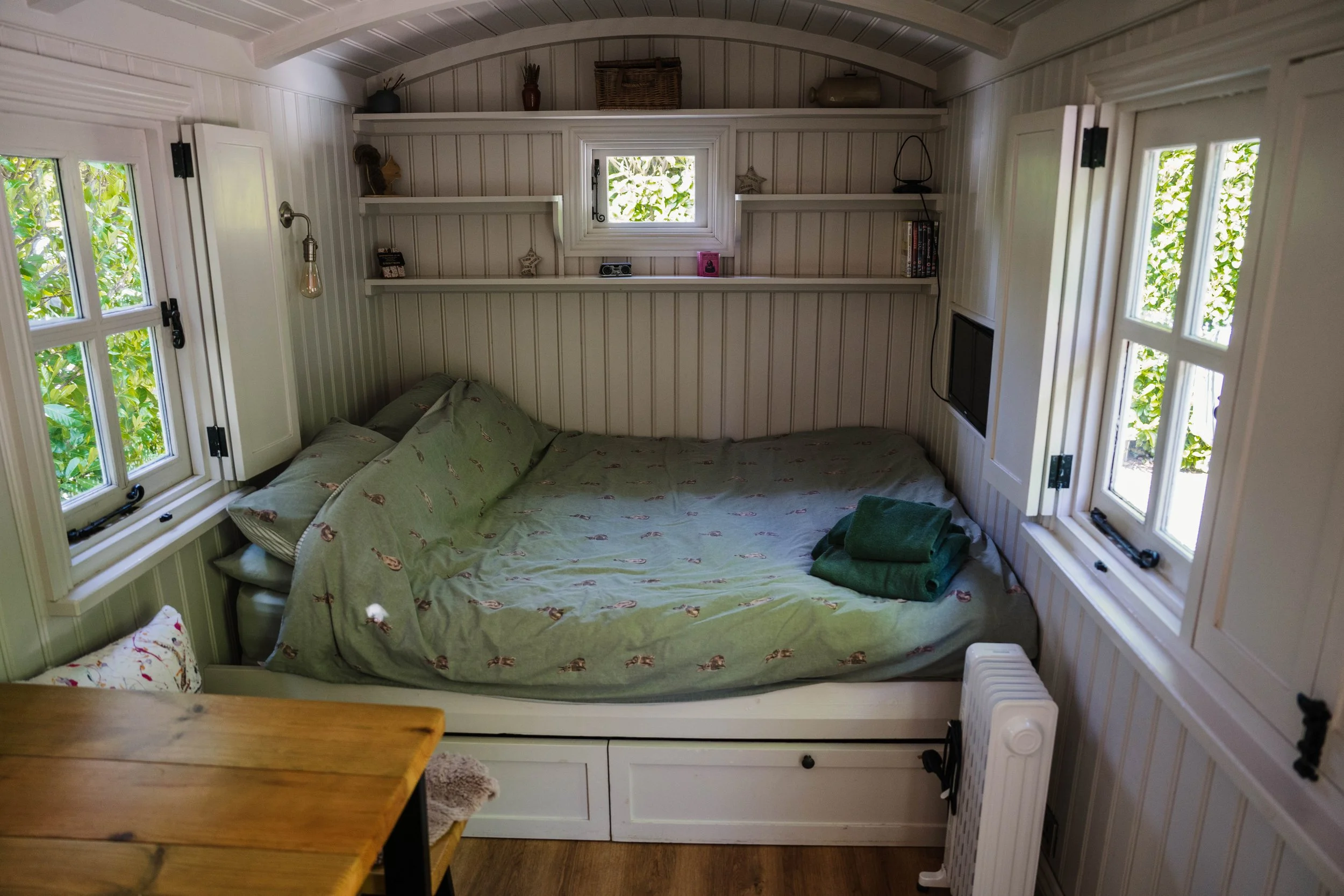 A cozy bedroom with white paneled walls and shelves. There are three windows with open shutters, allowing natural light in. The bed has green bedding with a printed pattern, and folded green towels are placed on top. There are decorative items on the shelves, including vases, books, and small ornaments. A TV is mounted on the wall near the bed, and a small white heater is visible at the bottom right corner.