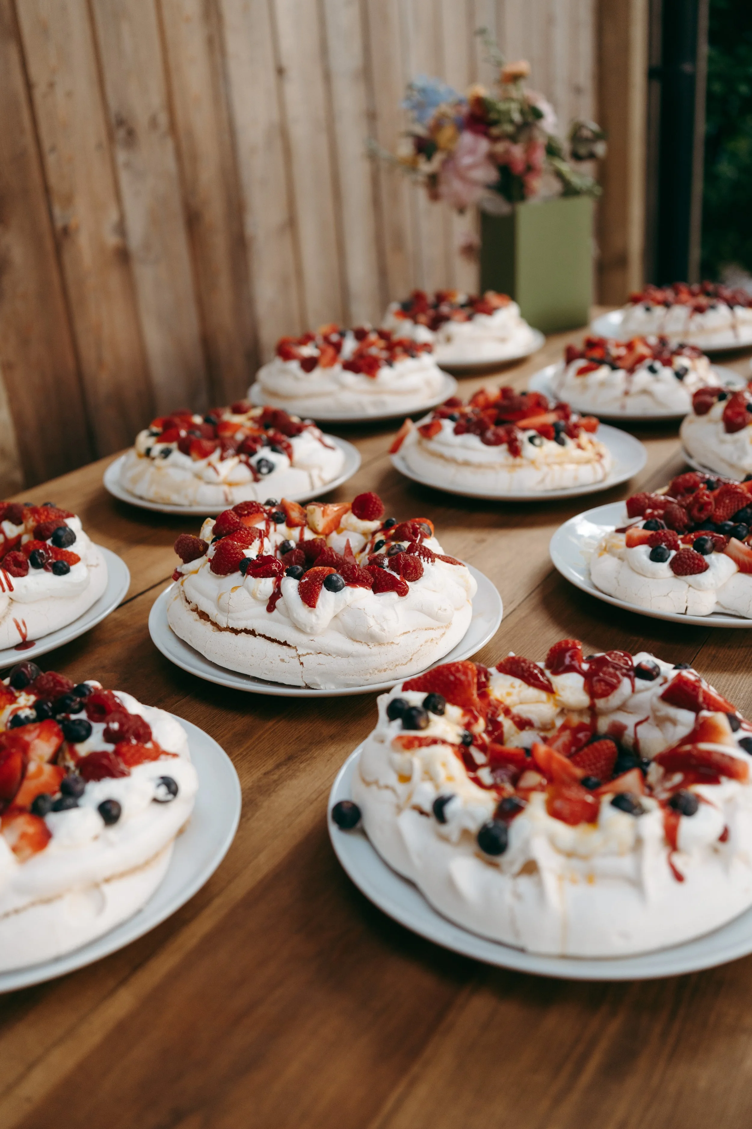 Assorted pavlova desserts topped with fresh berries on white plates on a wooden table with a flower arrangement in the background.