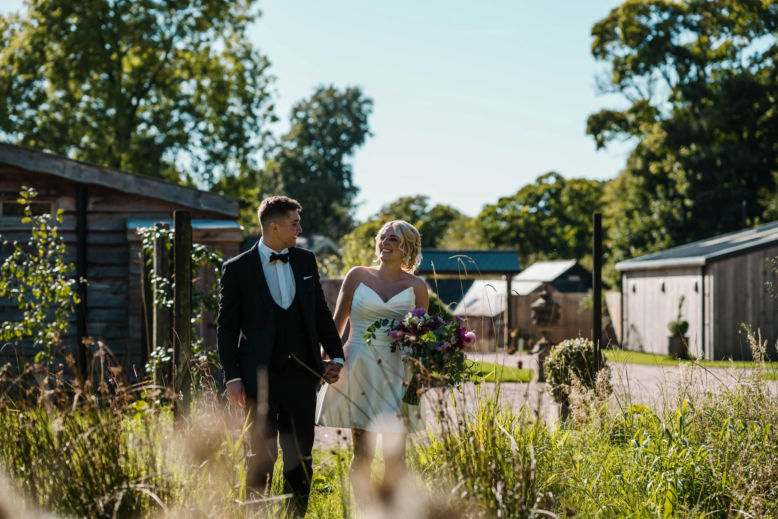 A newlywed couple walking outdoors in a rural setting, holding hands, with trees and farm buildings in the background on a sunny day.