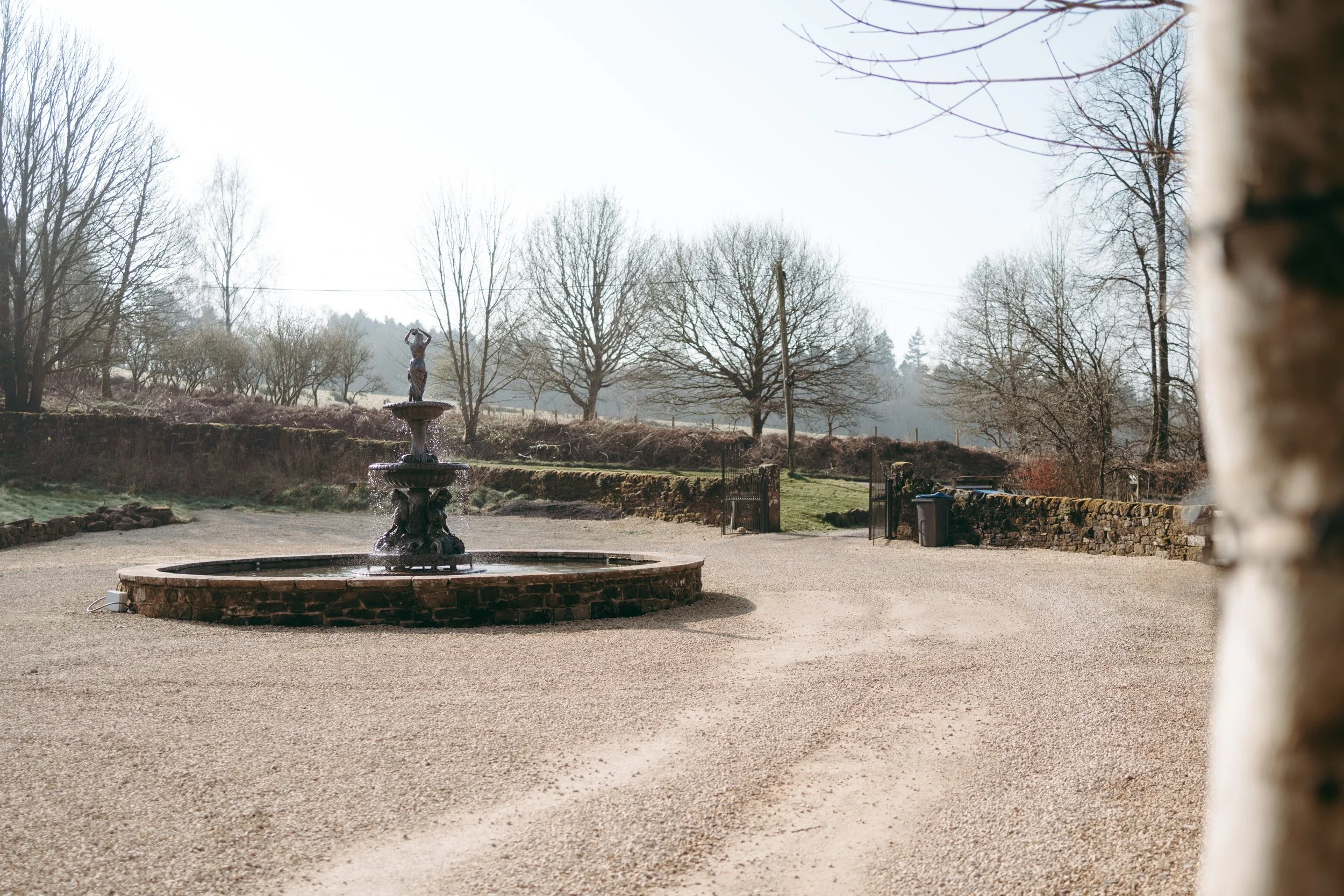 A stone fountain with water and a sculpture on top, located in an outdoor area with trees and a stone wall in the background, on a bright day.