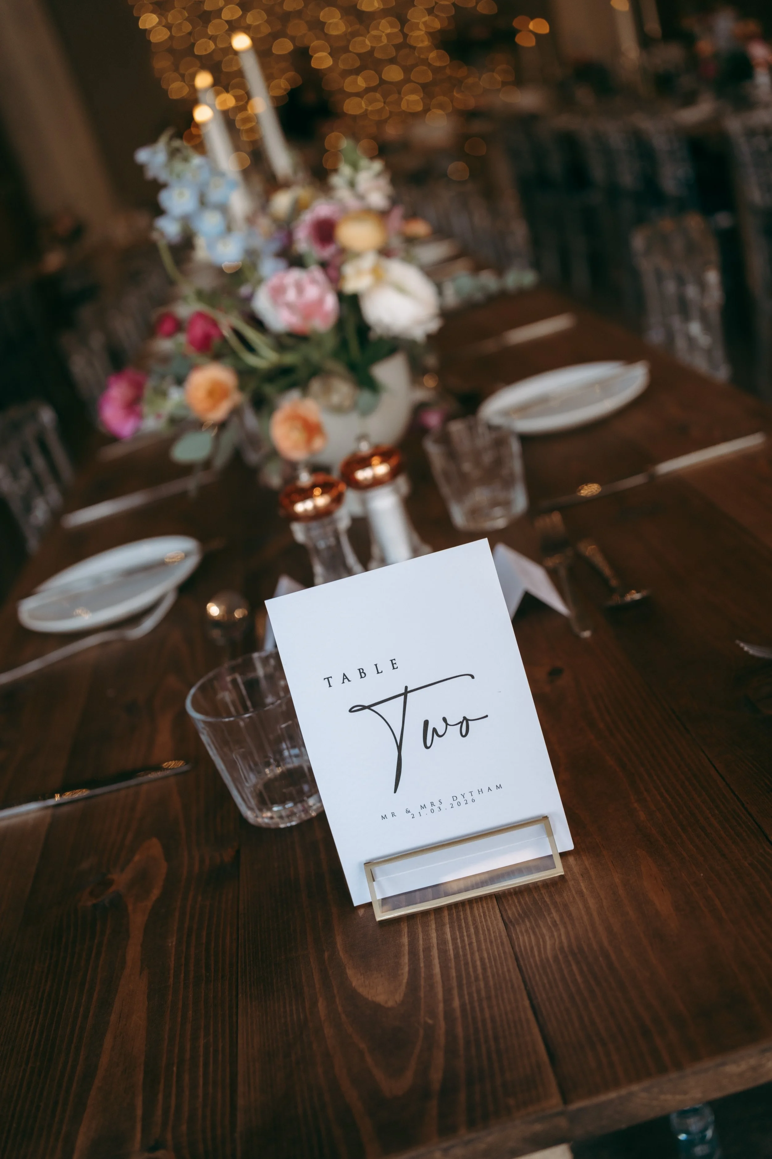 A beautifully decorated formal dining table with a centerpiece of colorful flowers, candles, and multiple place settings. There is a table number card indicating this is table two, with the names Mr. & Mrs. Dytham and the date 21.03.2026.