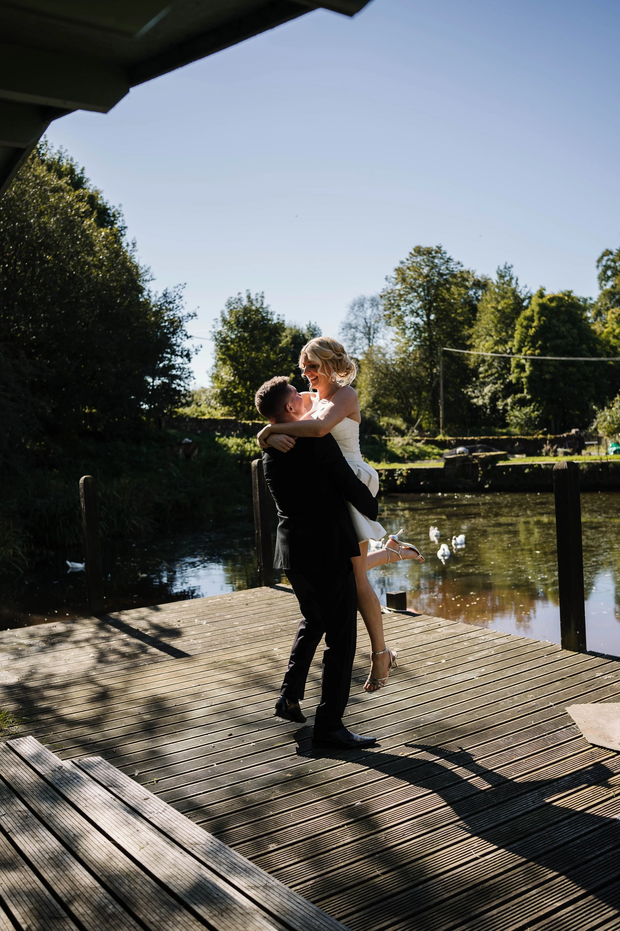 A couple dressed in wedding attire embracing each other on a wooden dock over a pond, with ducks swimming nearby, surrounded by trees under a clear blue sky.
