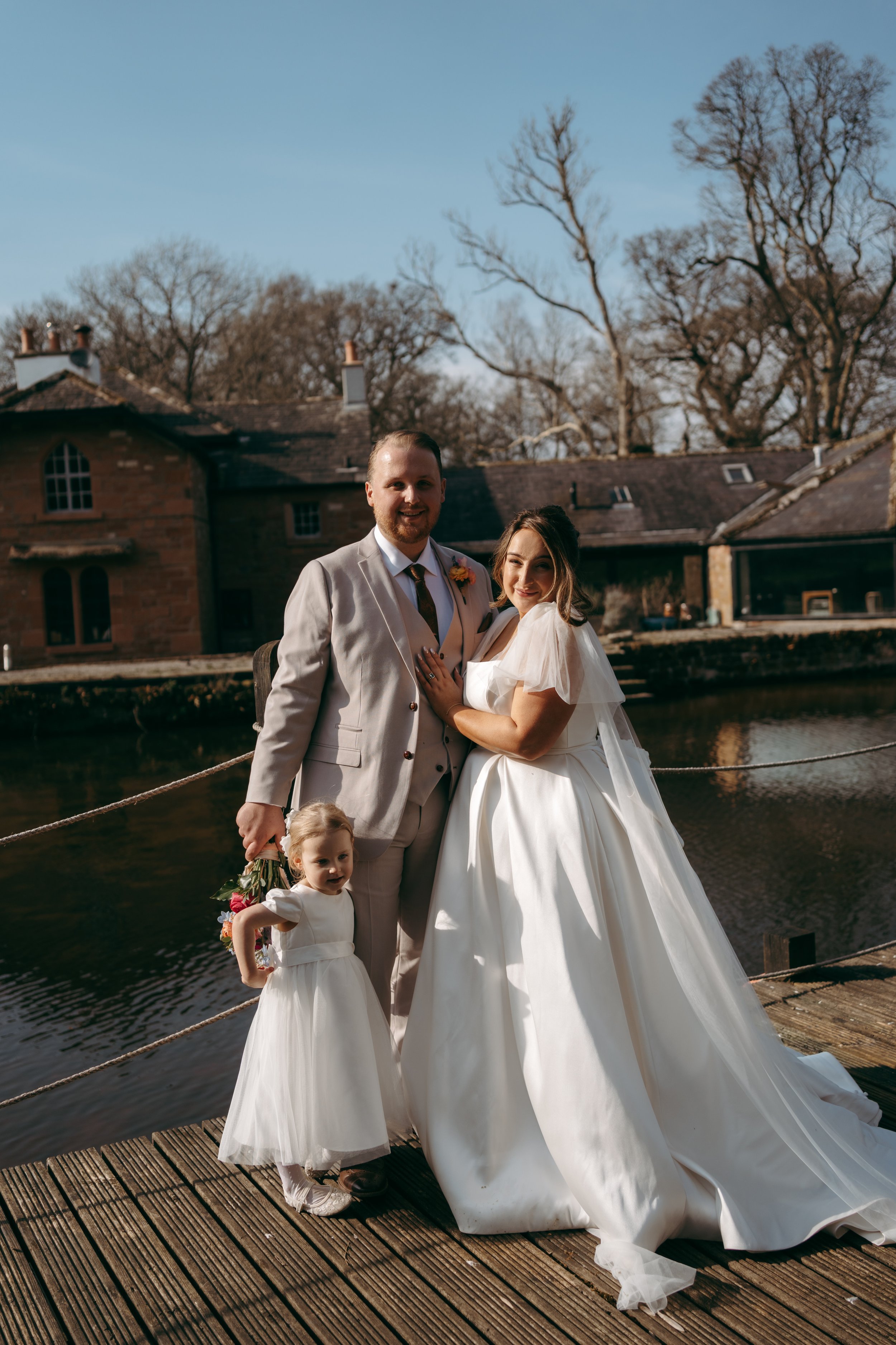 A bride and groom standing on a wooden dock near a river, with a young girl standing in front of them, all dressed in white. The bride is wearing a white wedding gown with puffed sleeves, and the groom is in a light-colored suit. The girl is holding 
