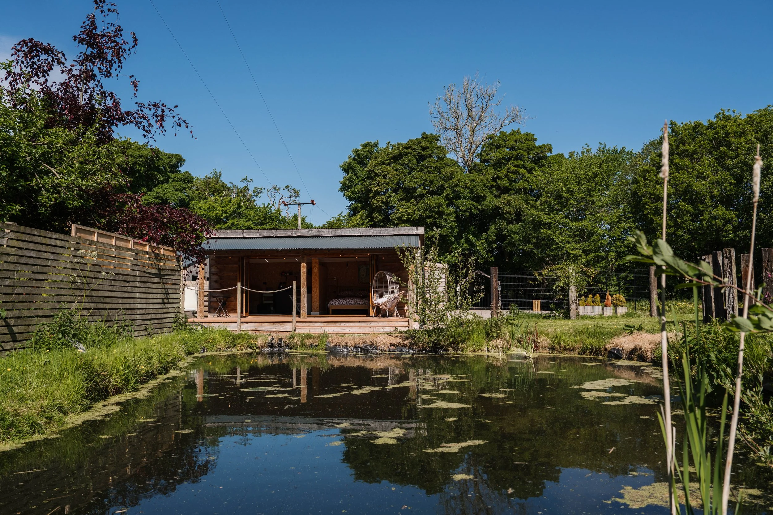 A backyard pond reflecting the blue sky and surrounding trees, with a small wooden outdoor living space and garden fence in the background.