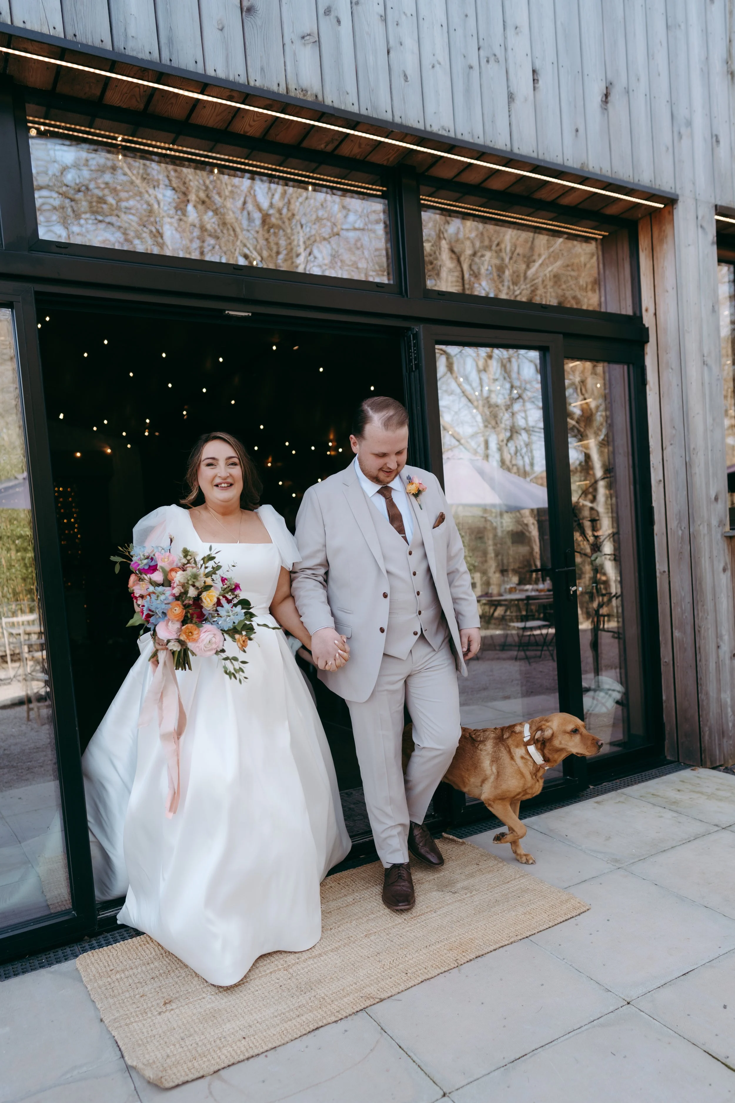 A bride and groom walking out of a building holding hands, accompanied by a brown dog on a leash. The bride is smiling and holding a bouquet of flowers, while the groom is looking down. They are dressed in wedding attire and are standing on a woven r