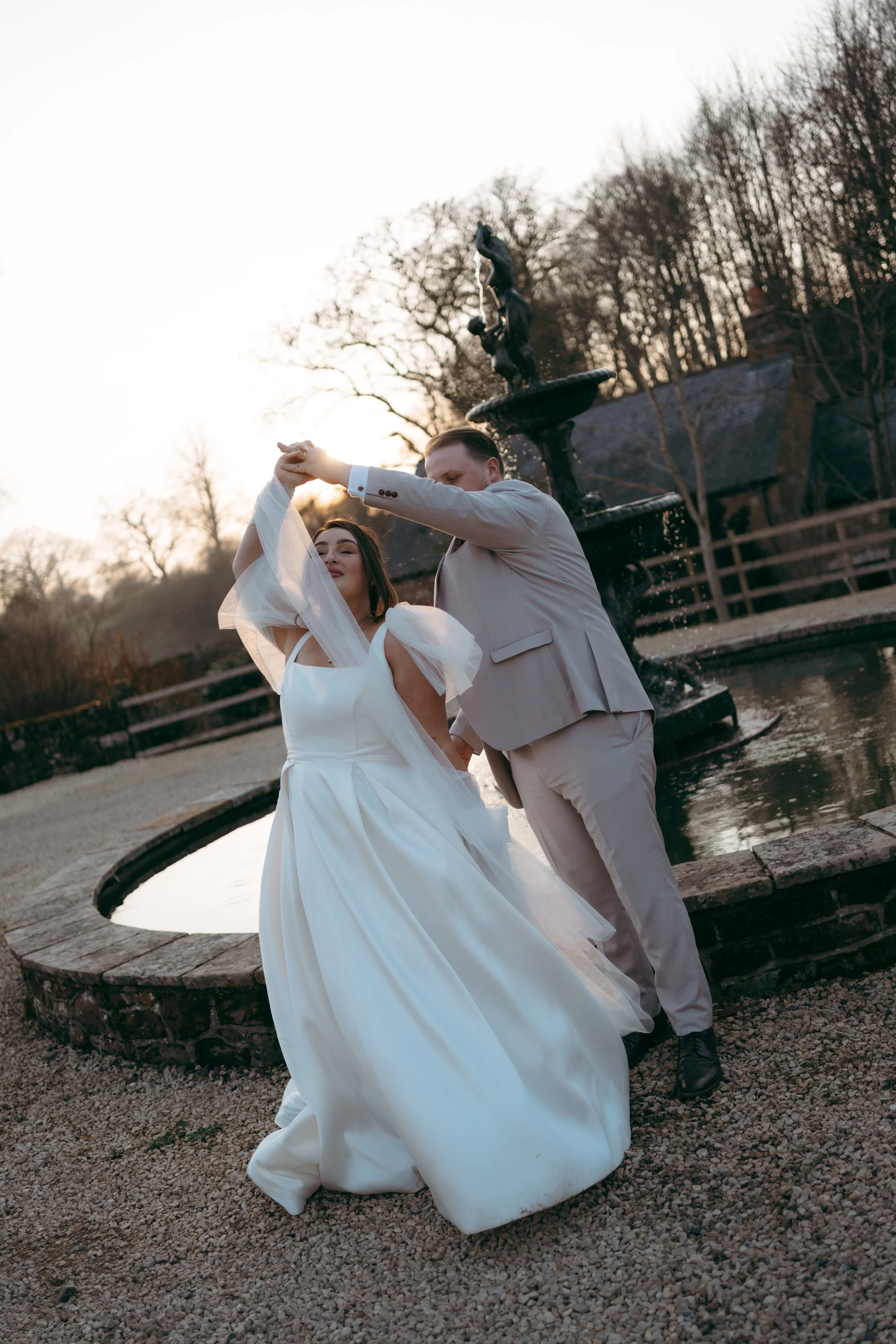 A couple dancing outdoors near a fountain with leafless trees in the background during sunset. The woman is wearing a white dress with sheer sleeves, and the man is dressed in a light beige suit.