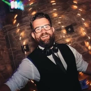 A smiling man with glasses and a beard, wearing a dress shirt and vest, standing in front of a brick wall with festive lighting, in a nightclub or party setting.