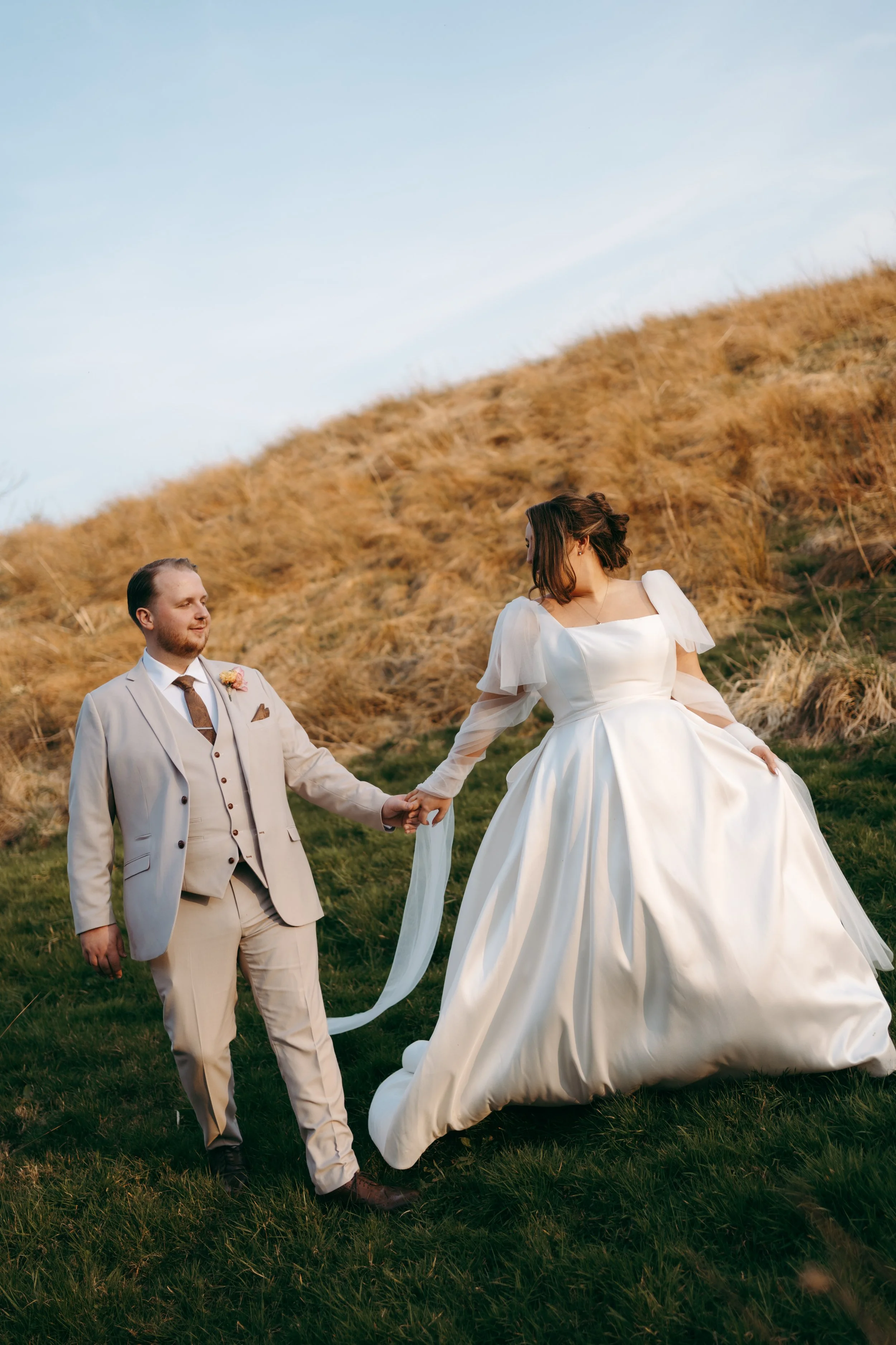 A man and woman in wedding attire holding hands in a grassy outdoor setting with a hill of dry grass behind them.