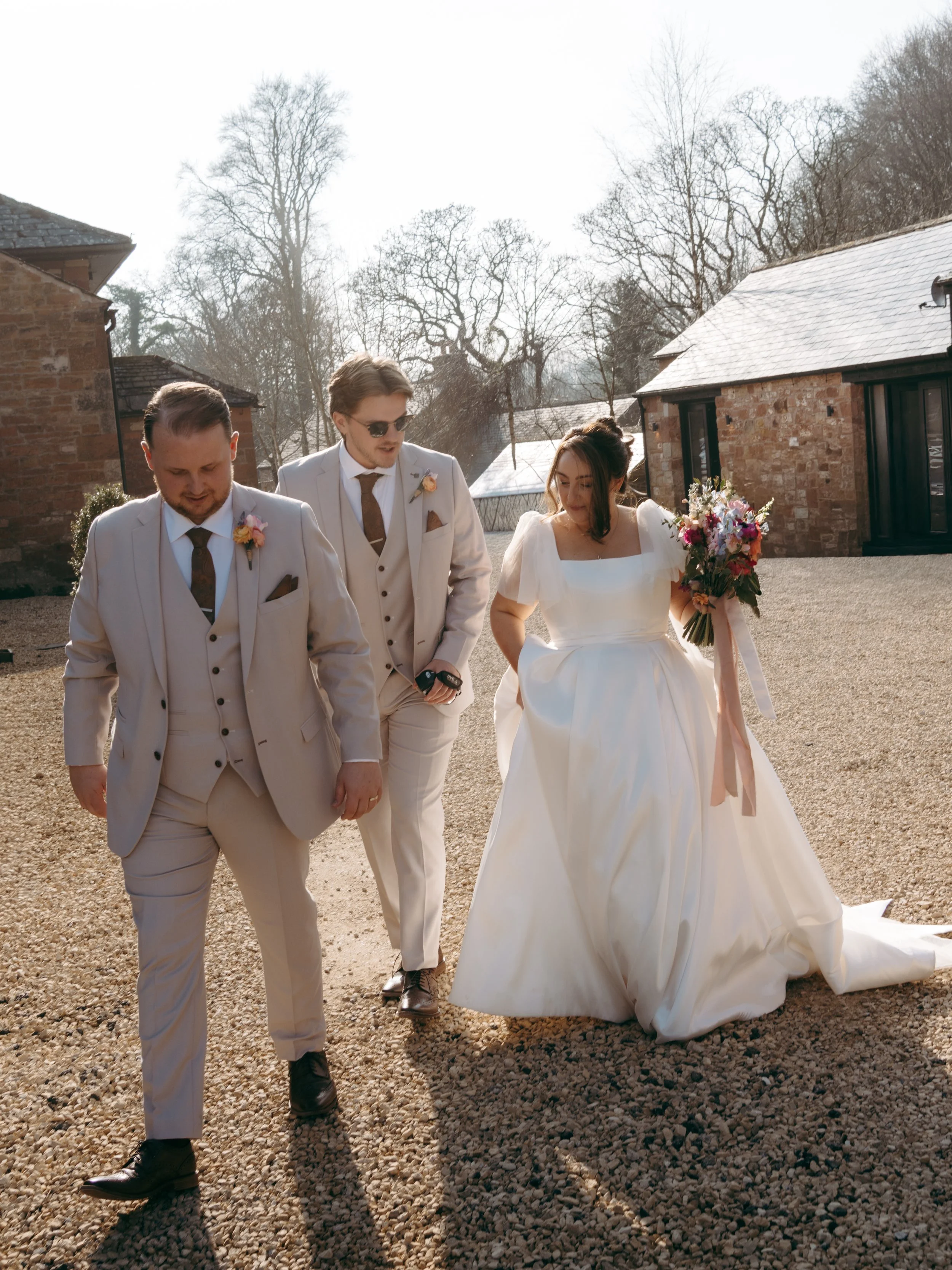 A bride and two groom are walking outside near brick buildings on a sunny day, dressed in wedding attire.