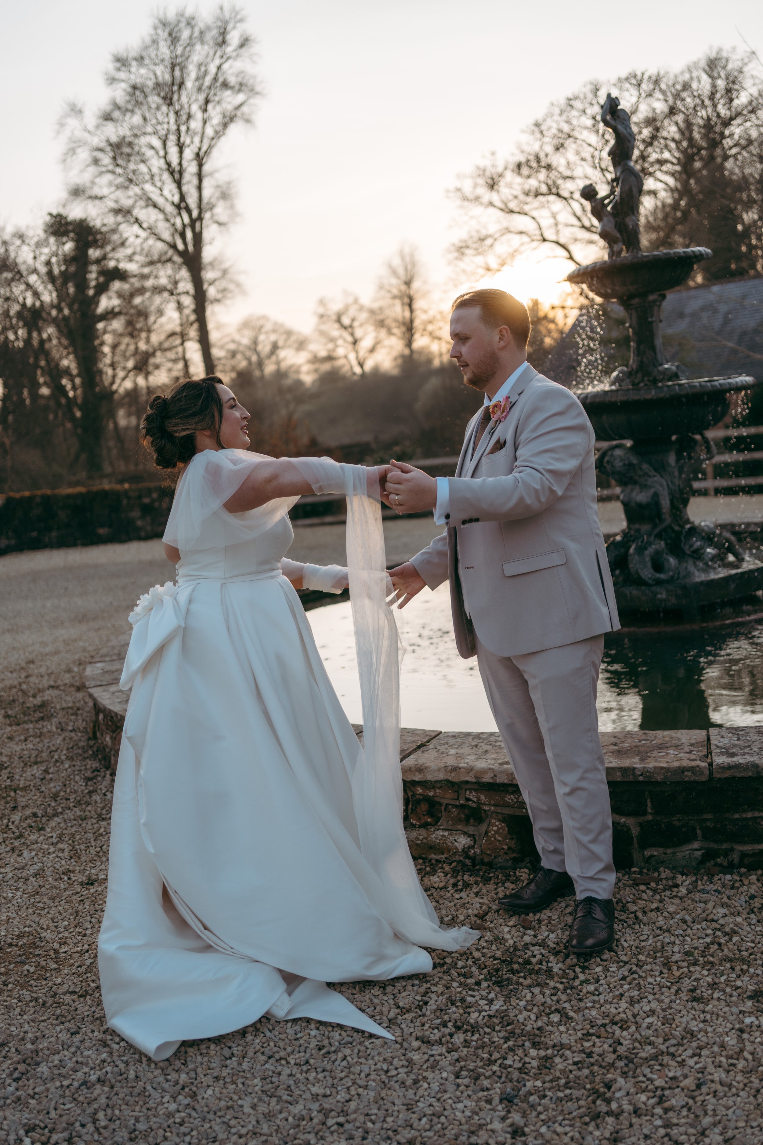 A bride and groom are holding hands and dancing outdoors near a fountain during sunset. The bride is wearing a white wedding gown and the groom is dressed in a light-colored suit with a flower boutonniere. There are leafless trees in the background.