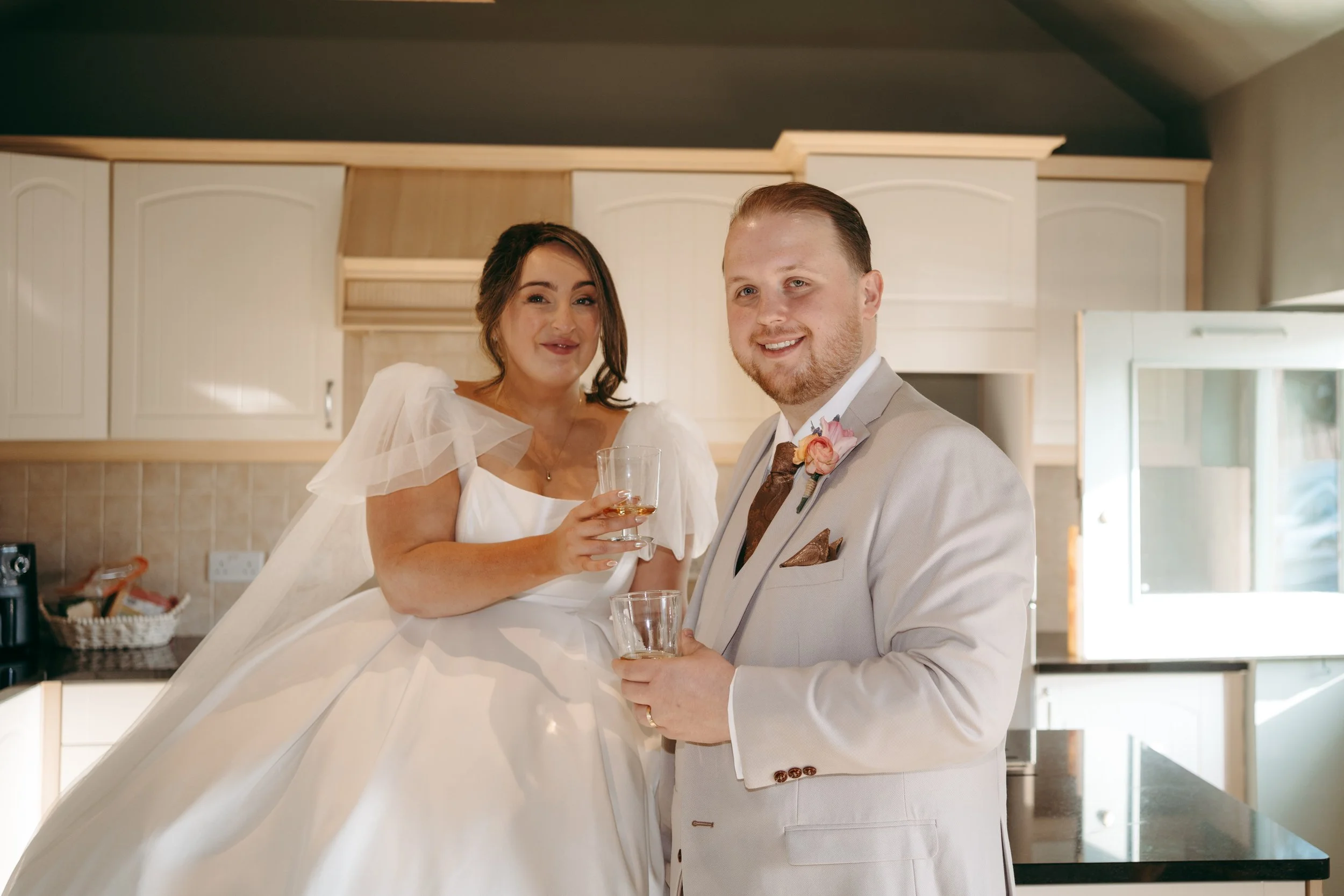 A bride and groom smiling with drinks in hand in a kitchen.
