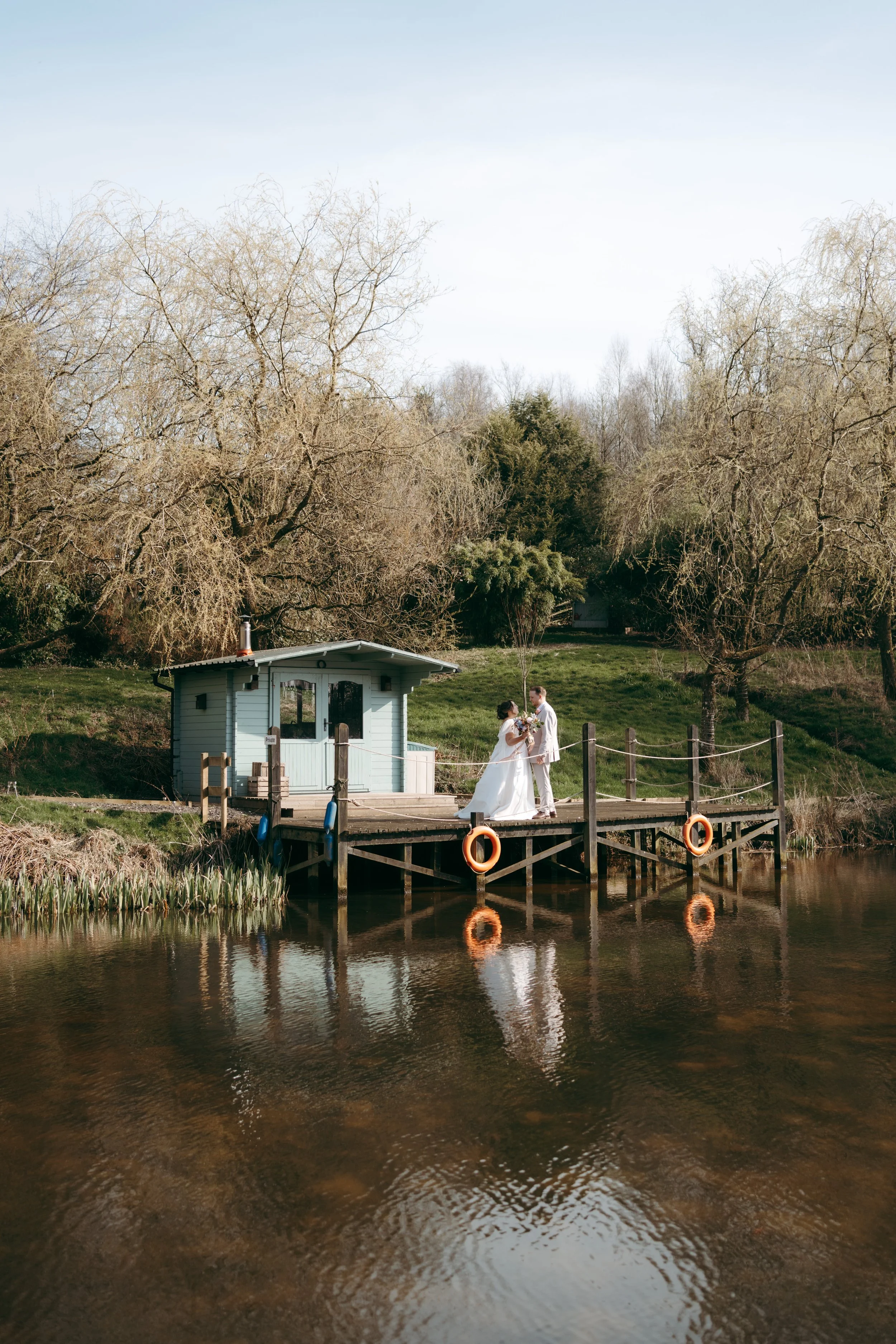 A bride and groom in wedding attire standing on a wooden dock beside a small white house by a pond, with barren trees in the background and their reflection in the water.