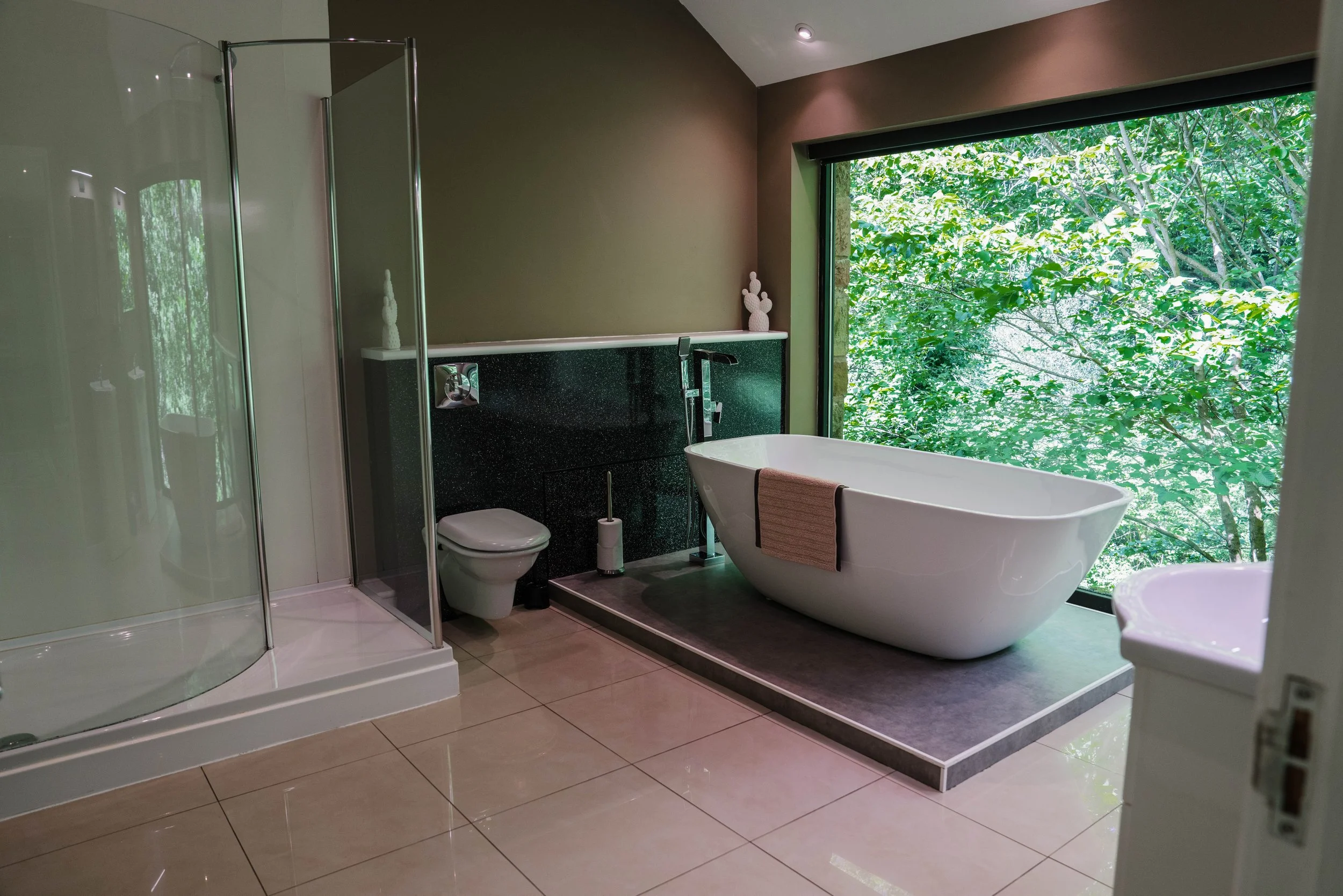 Modern bathroom with a freestanding bathtub near a large window showing lush green trees outside, a white toilet, a glass shower enclosure, and minimal decorative elements.