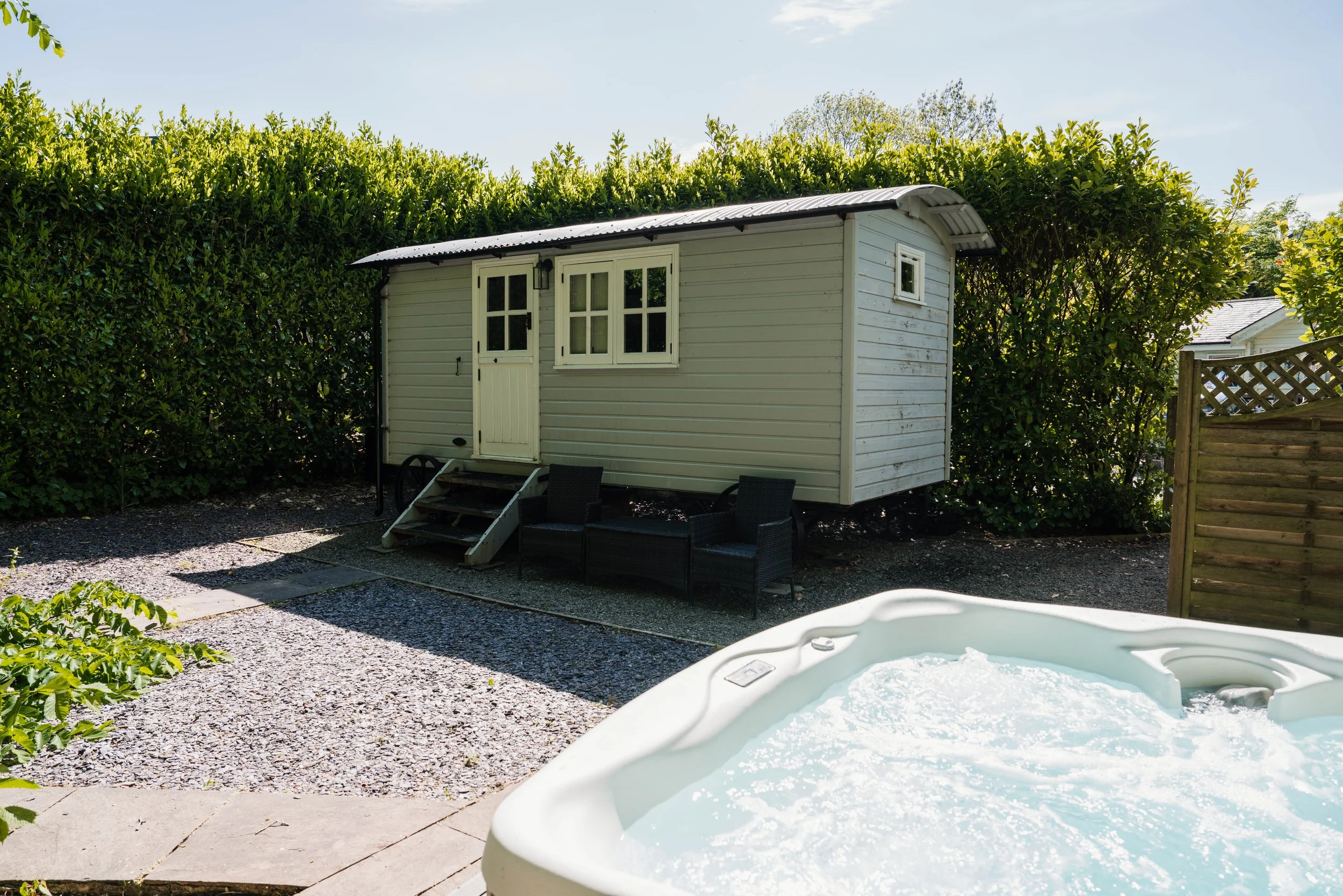A small white shed with a curved roof, set against a hedge and a blue sky; outdoor furniture and a hot tub with bubbling water are visible in the yard.