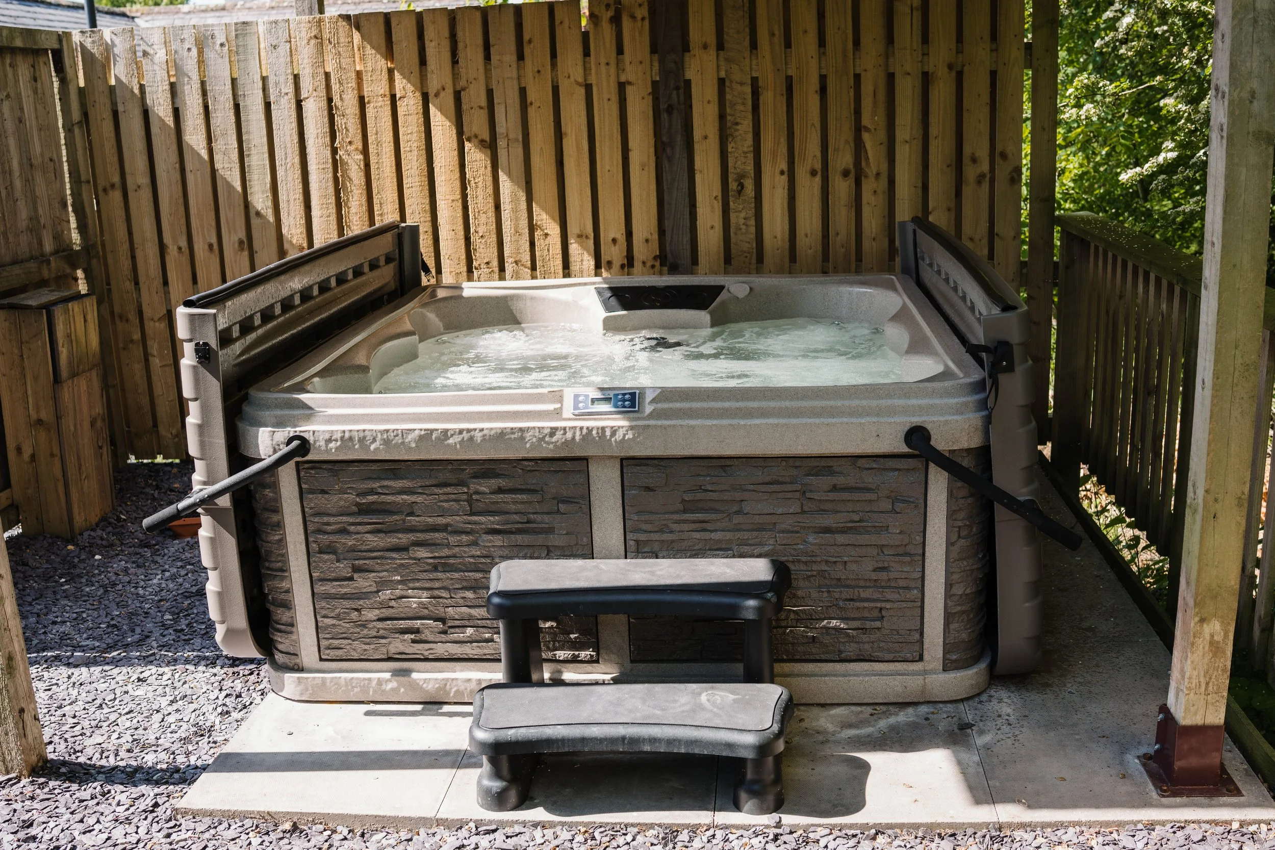 An outdoor hot tub with bubbling water, placed on a concrete pad, with a wooden privacy fence around it and a small black step stool in front.