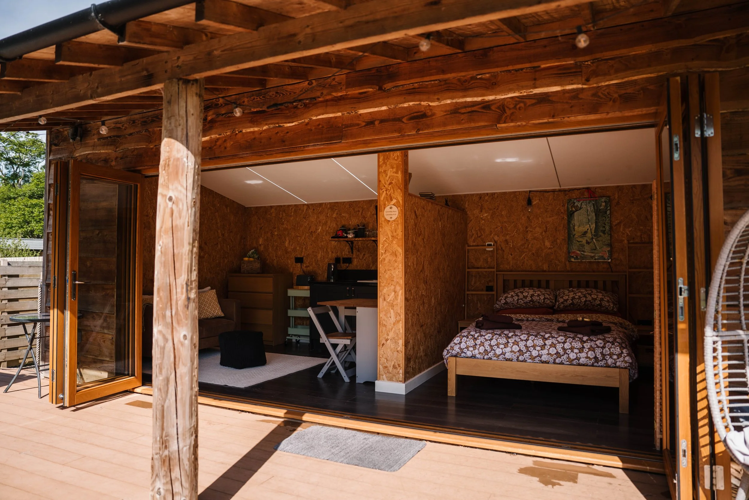 An open wooden deck with a view into a cozy bedroom with wooden walls, a bed with floral sheets, a small window, and interior furniture.